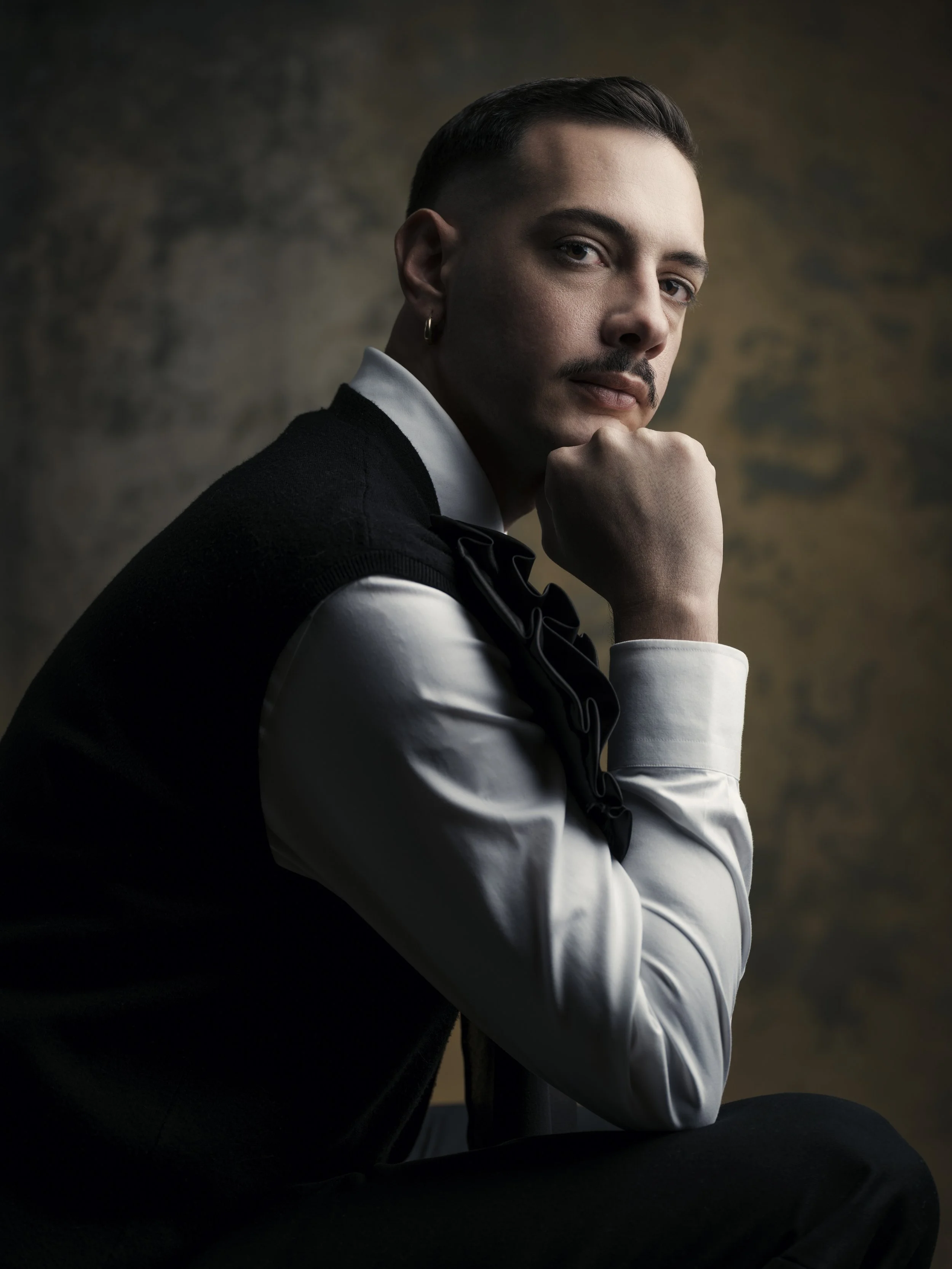 A man with dark hair and a mustache, wearing a white shirt, black vest, and a black bow tie, is sitting with his chin resting on his hand, looking at the camera, against a dark, textured background.