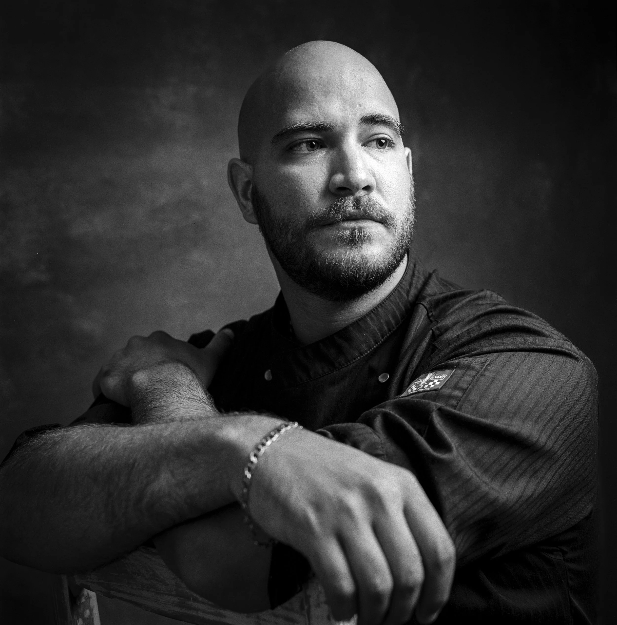 Black and white image of a man's face with a shaved head, beard, and mustache, looking off to the right, with a dark background.