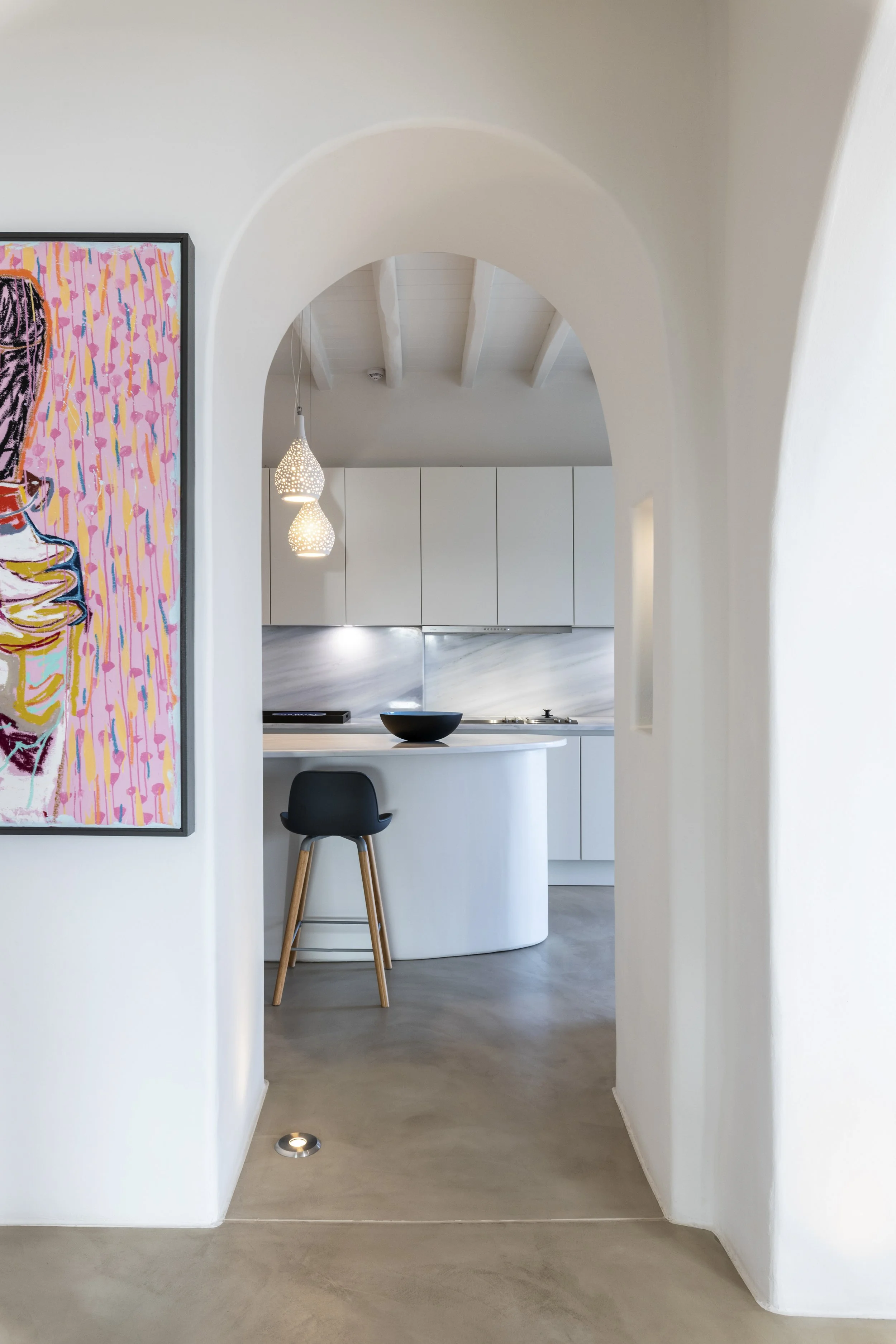 A modern minimalist kitchen viewed through an arched doorway, featuring white cabinets, pendant lighting, and a kitchen island with a black bowl and a black chair.
