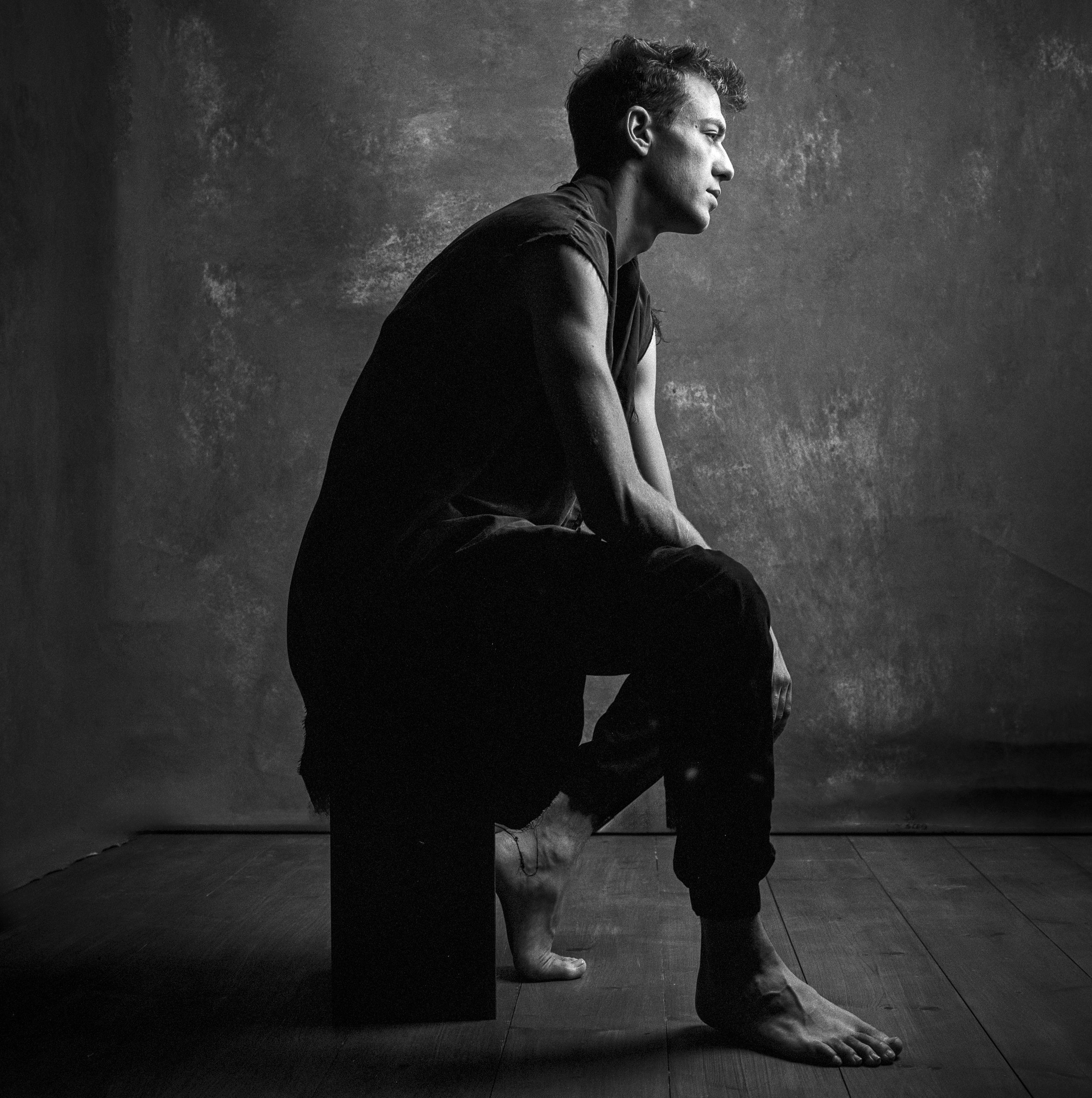 A black and white photo of a person in a yoga pose on a wooden floor against a textured background. The person is sitting with their legs crossed, leaning forward with one hand on the floor and the other hand resting on the knee, gazing to the side.