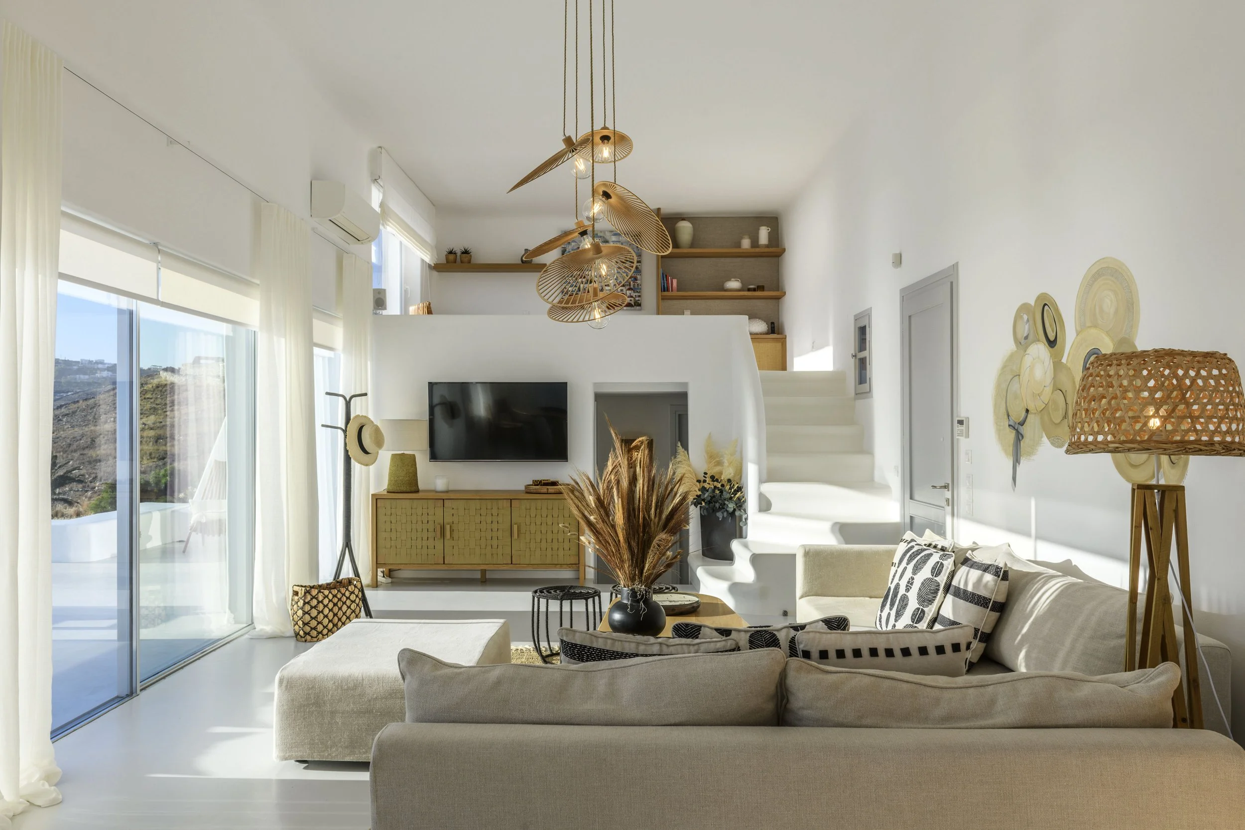 Bright, modern living room with large glass sliding doors, white walls, and ceiling. Interior decor includes a flat-screen TV, patterned black-and-white pillows on a cream sofa, wicker lampshade, and wall-mounted hats. Wooden shelves hold decorative 