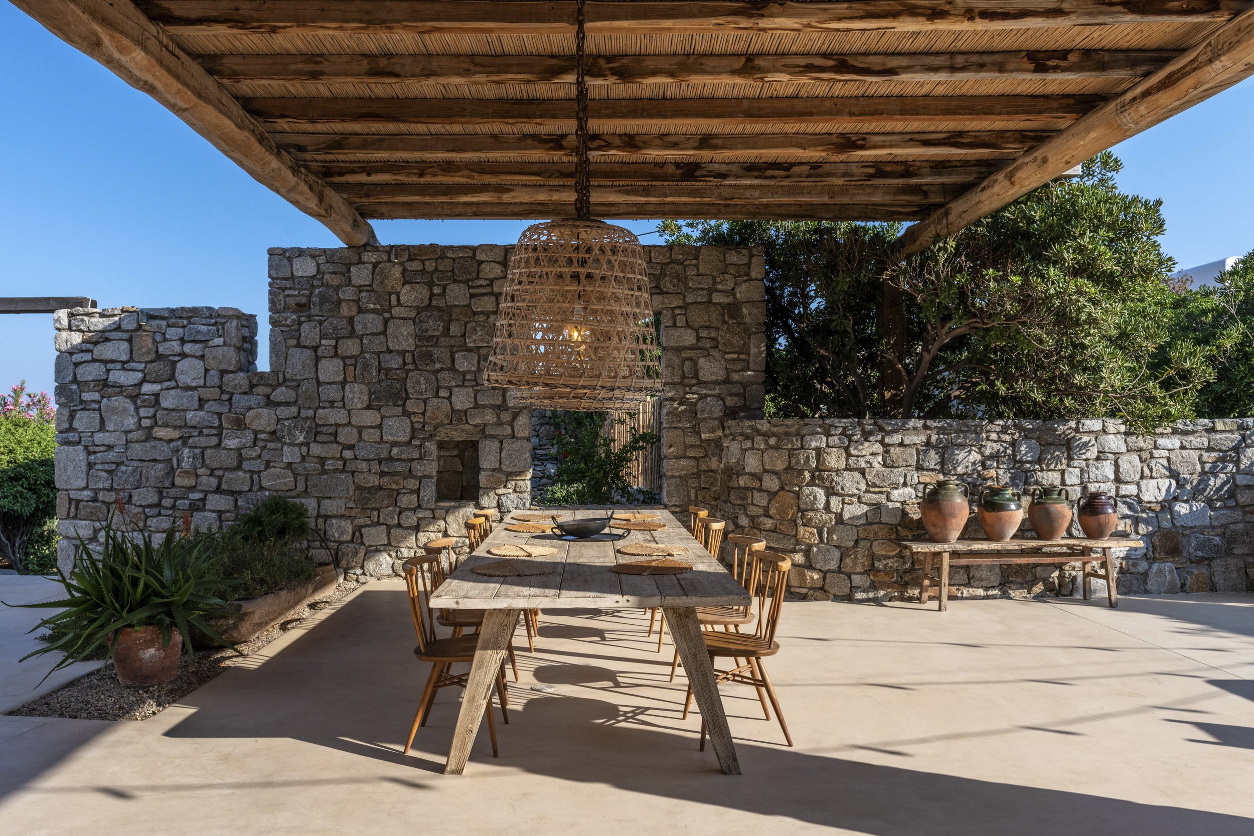 Outdoor dining area with a wooden table and chairs, stone walls, potted plants, and a woven pendant light.