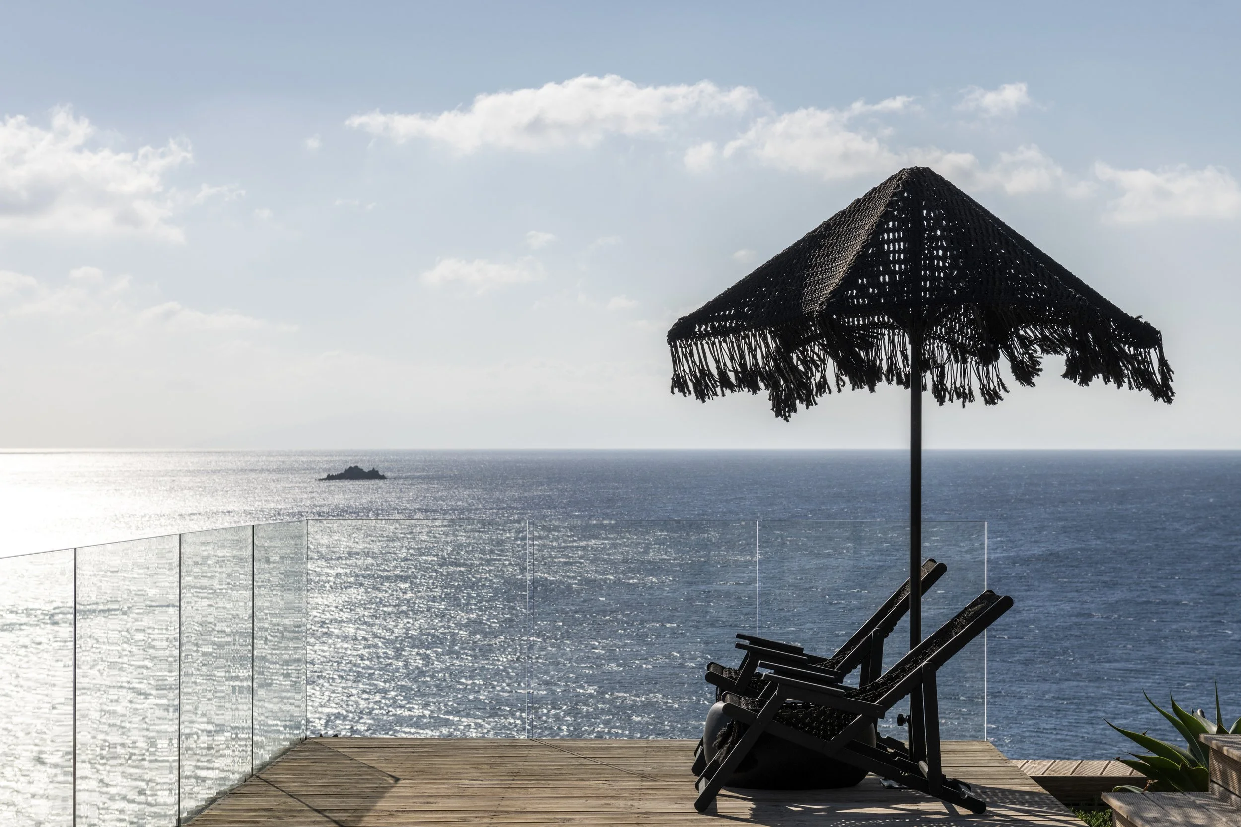 A patio with a wooden deck overlooking the ocean, with two lounge chairs and a large black straw beach umbrella, against a backdrop of blue sky with clouds.