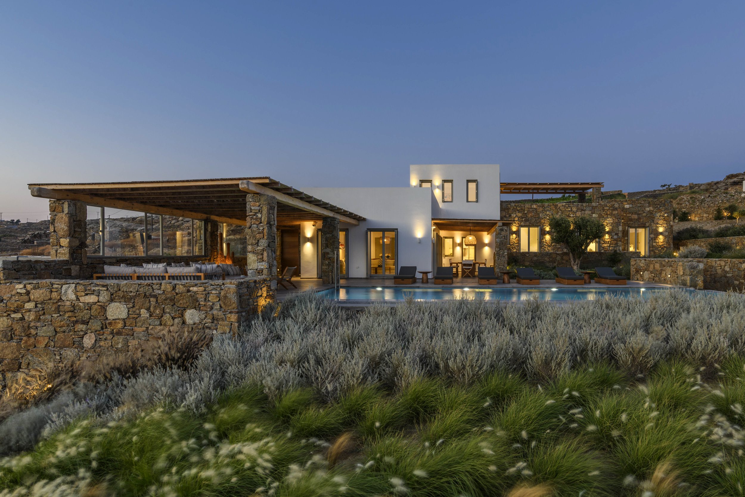 Modern house with stone and white exterior, large windows, outdoor seating area, pool, sunset lighting, surrounded by shrubs.