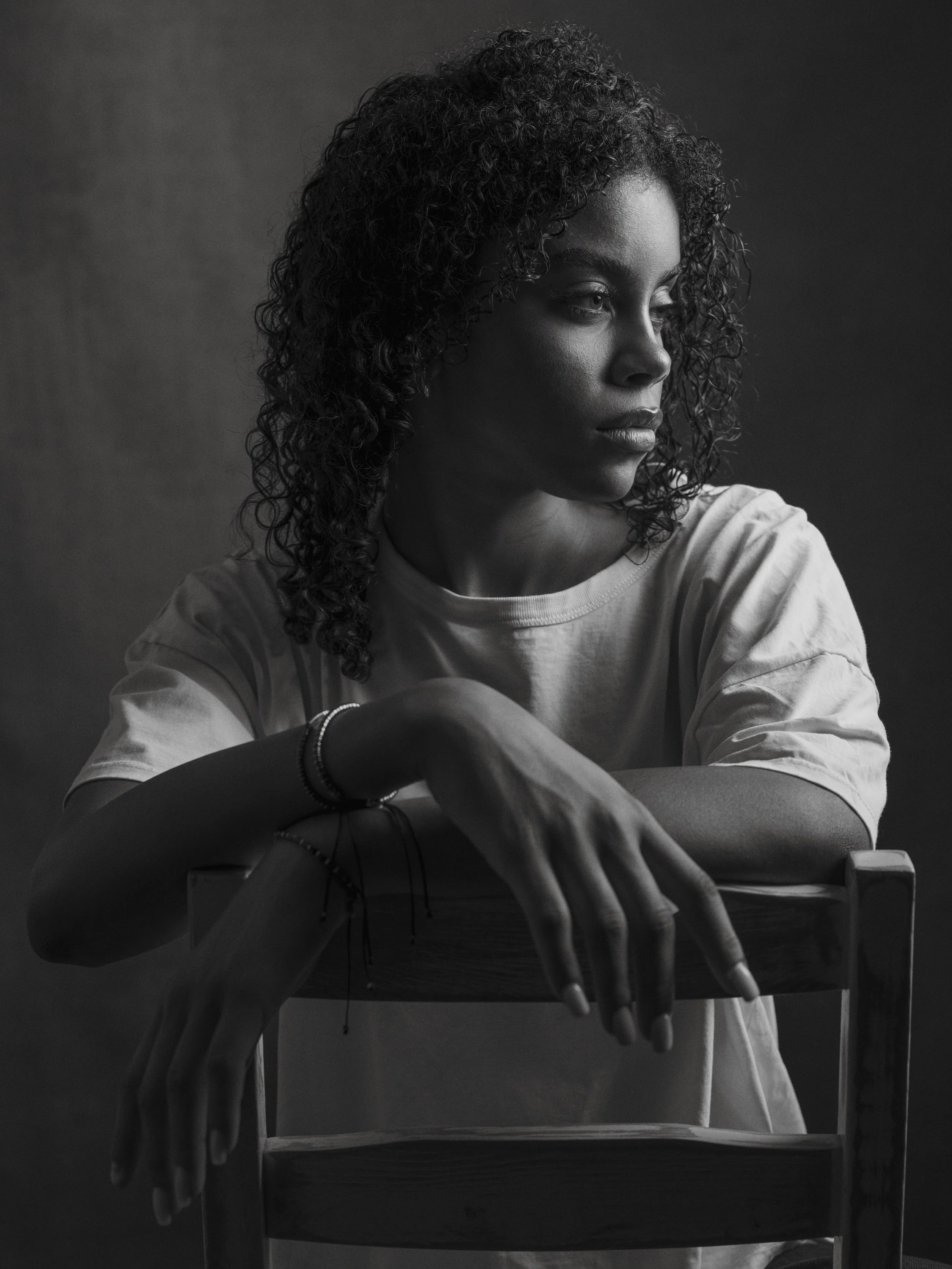 A black and white portrait of a young woman with curly hair sitting at a desk, resting her arms on the back of a chair in front of her, looking thoughtfully to the side.