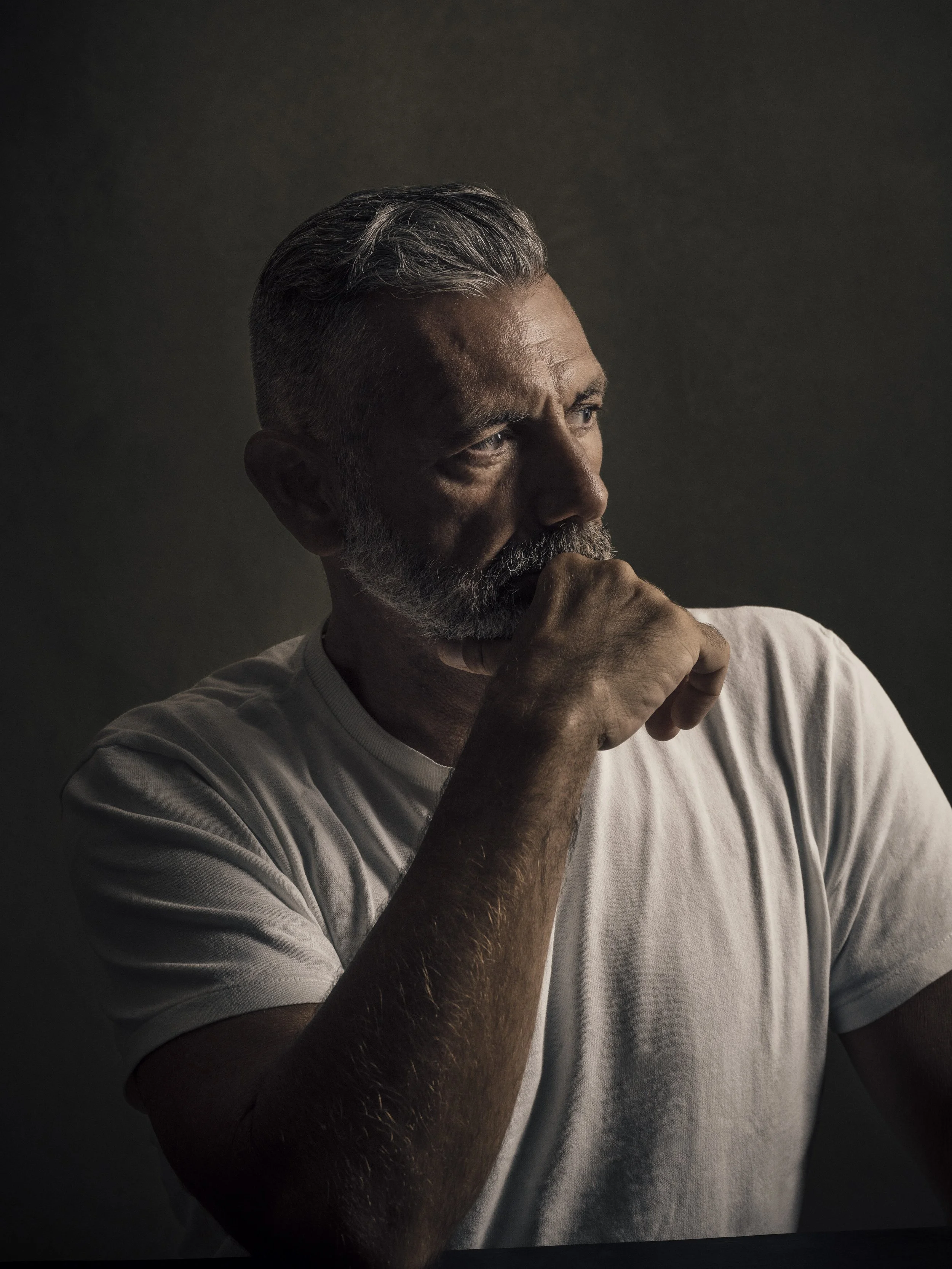 Close-up of a middle-aged man with gray hair and beard, wearing a white t-shirt, with his hand resting on his chin, looking thoughtfully to the right in a dimly lit setting.