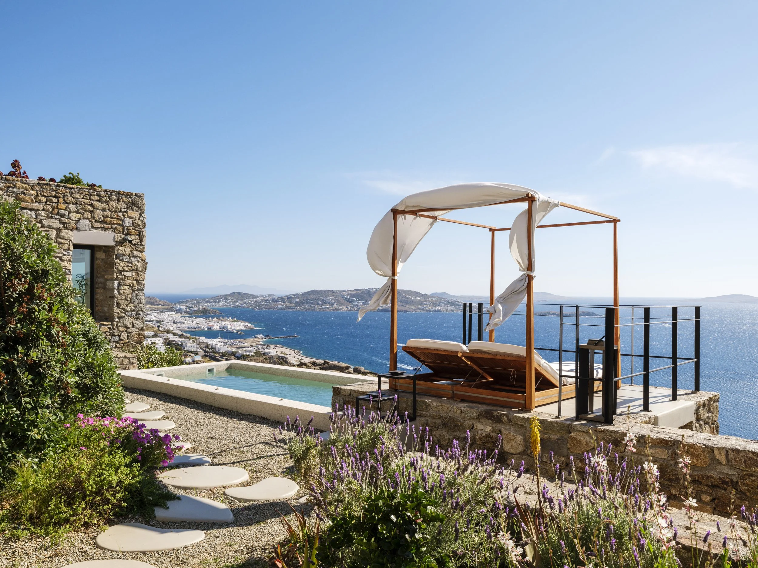 A scenic view of a coastal area with a stone building, colorful flowers, a swimming pool, and a wooden canopy with white fabric, overlooking the ocean and distant islands under a clear blue sky.