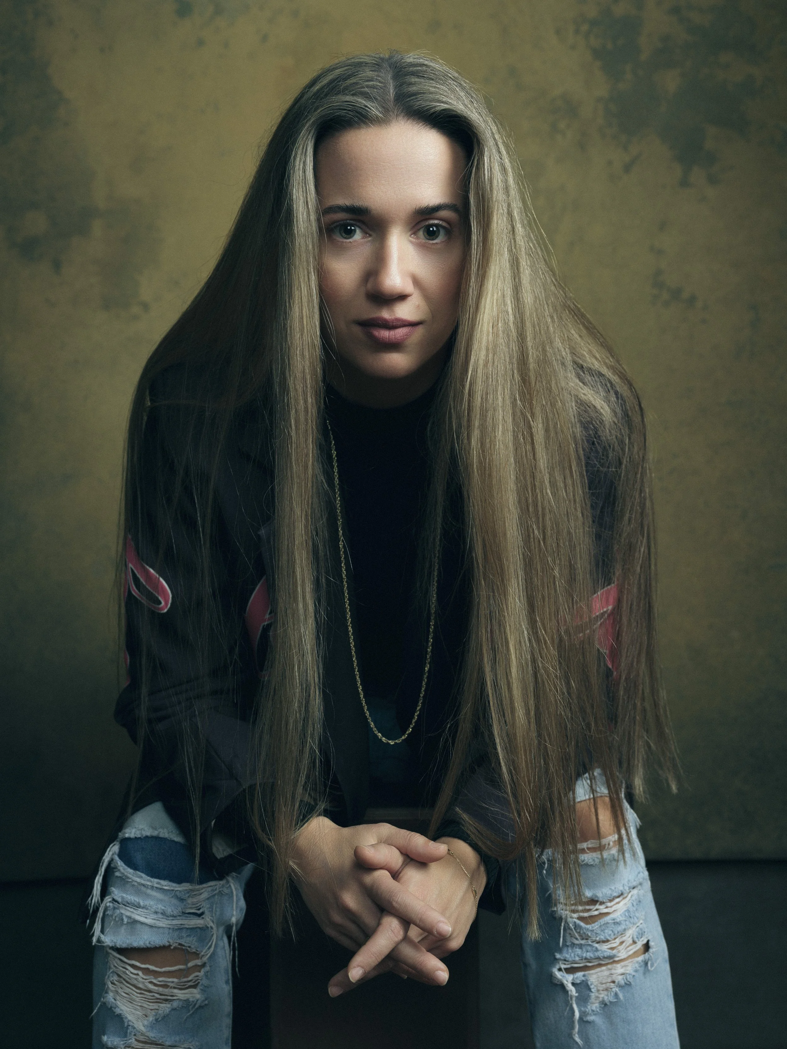 A young woman with long blonde hair, wearing a black top and distressed jeans with rips, sitting with her hands clasped in front of her against a dark background.