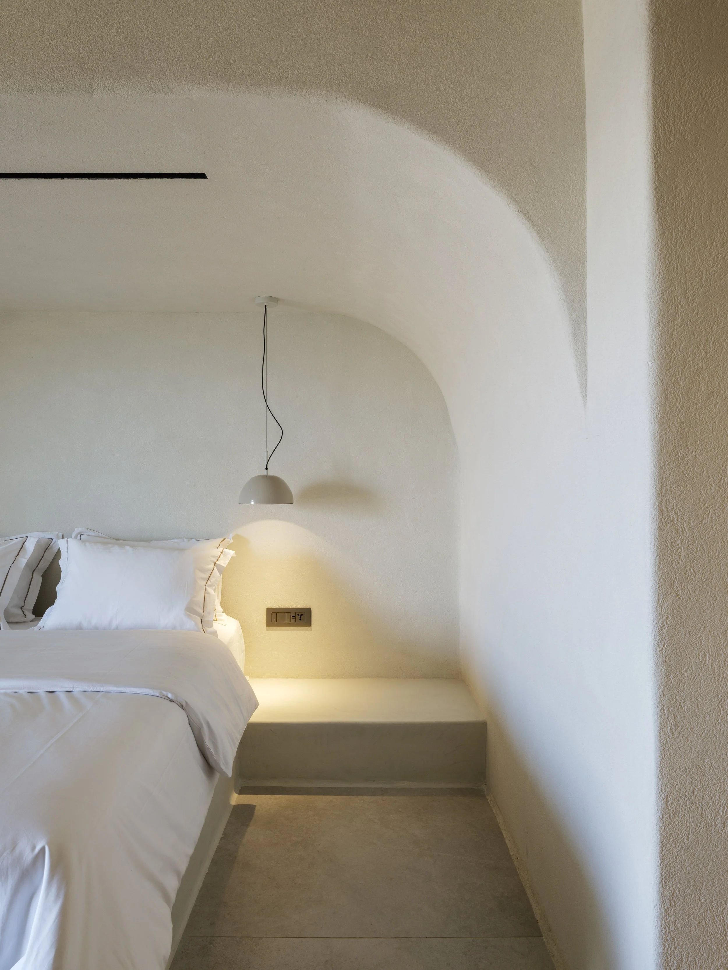 A bedroom with white bedding, a pendant light hanging from the ceiling, and a wall socket with switches near the bed.