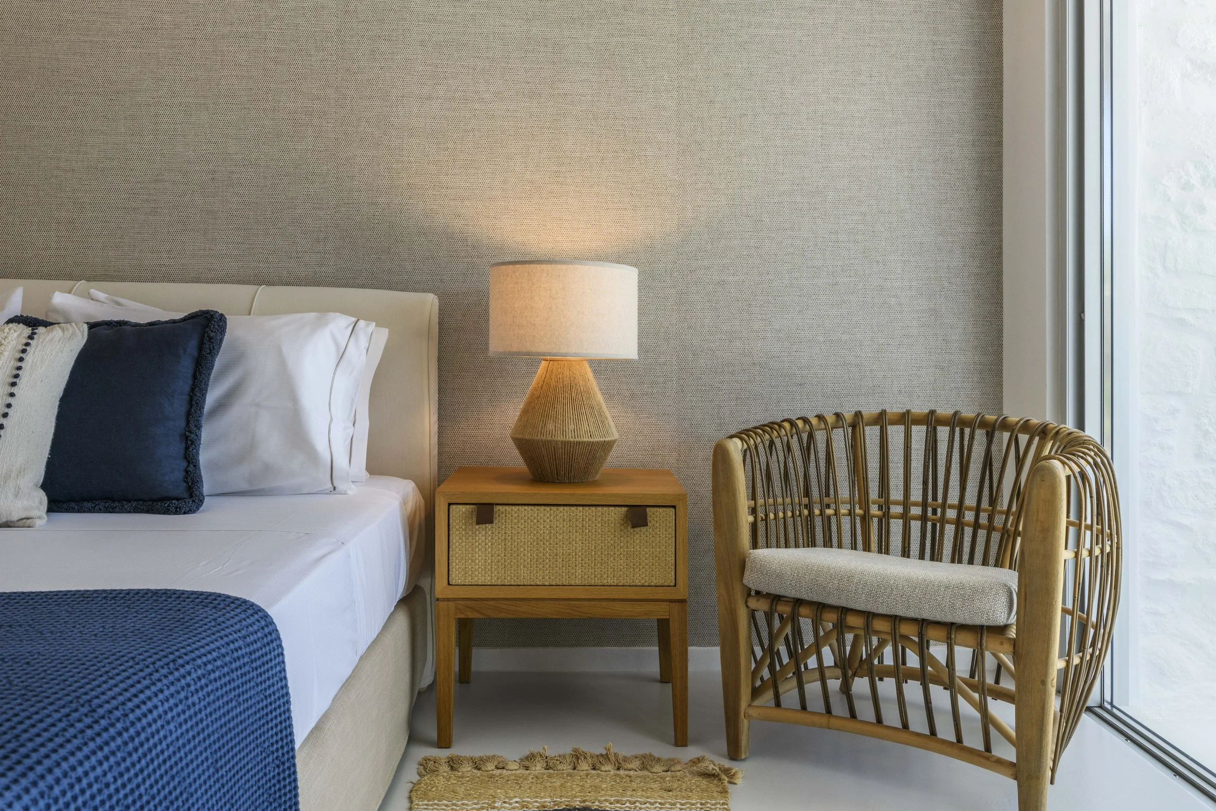 A bedroom with beige walls, a bed with white linens and navy cushions, a wooden nightstand with a woven lamp, and a wooden chair with a woven backrest. A window with white trim is on the right.