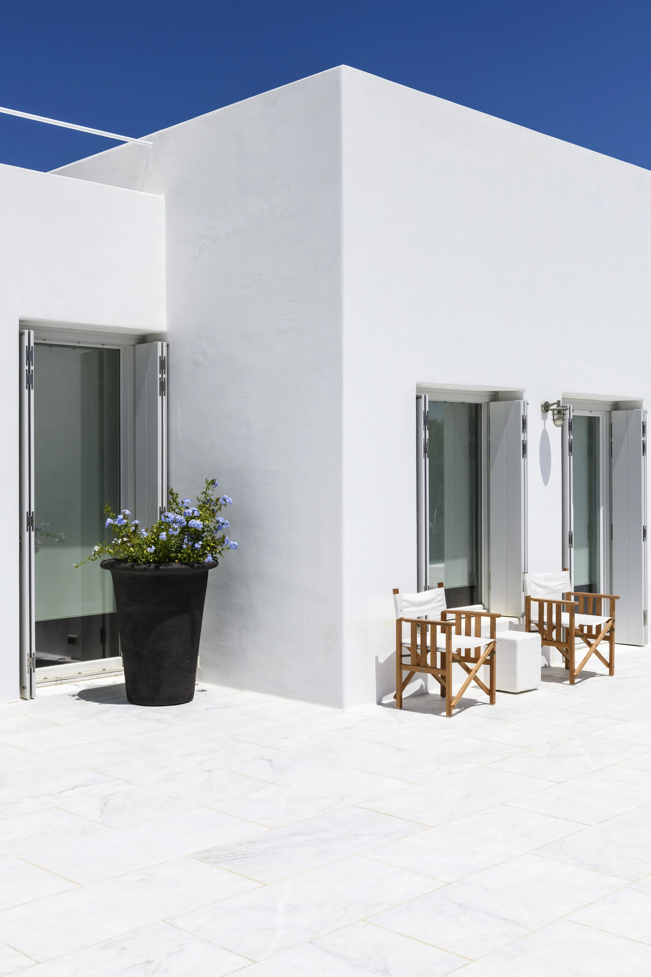 White building with open shutters, outdoor seating with wooden chairs, potted plant with purple flowers, blue sky.