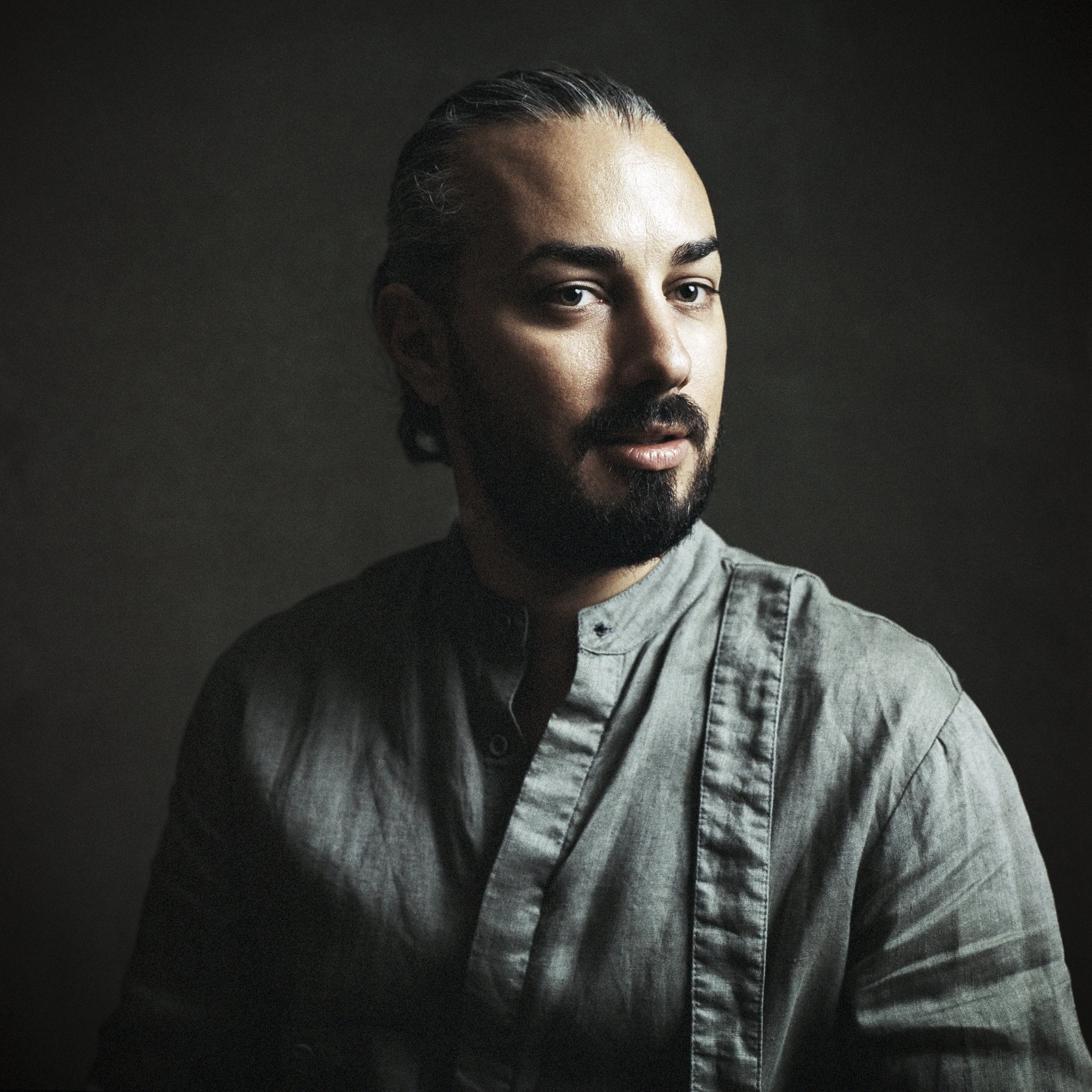 Close-up of a man with a beard and mustache, wearing a gray shirt, against a dark background.