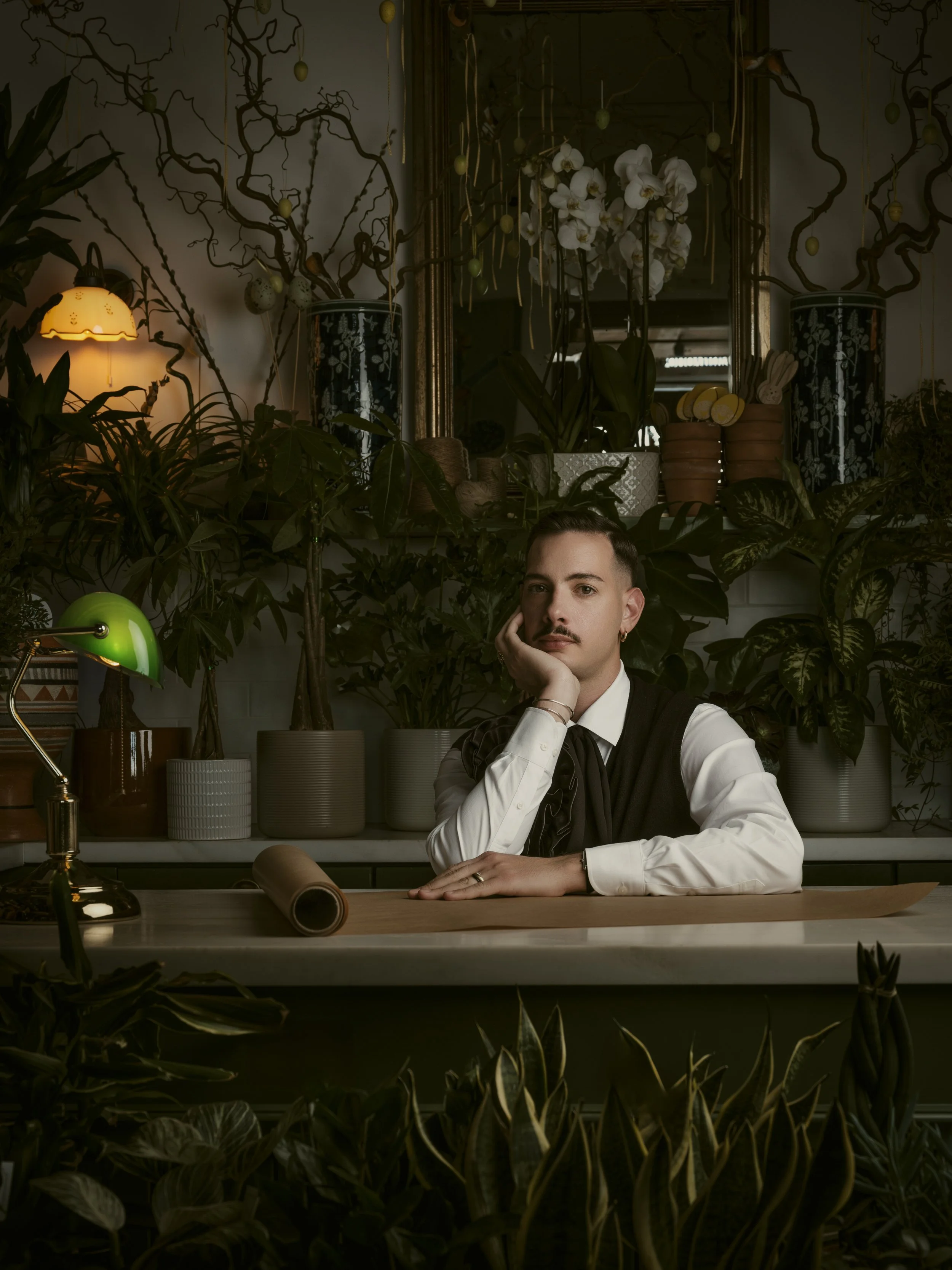 A man with short dark hair, a mustache, and earrings, sitting at a desk surrounded by potted plants, with a mirror, flowers, and decorative items in the background.