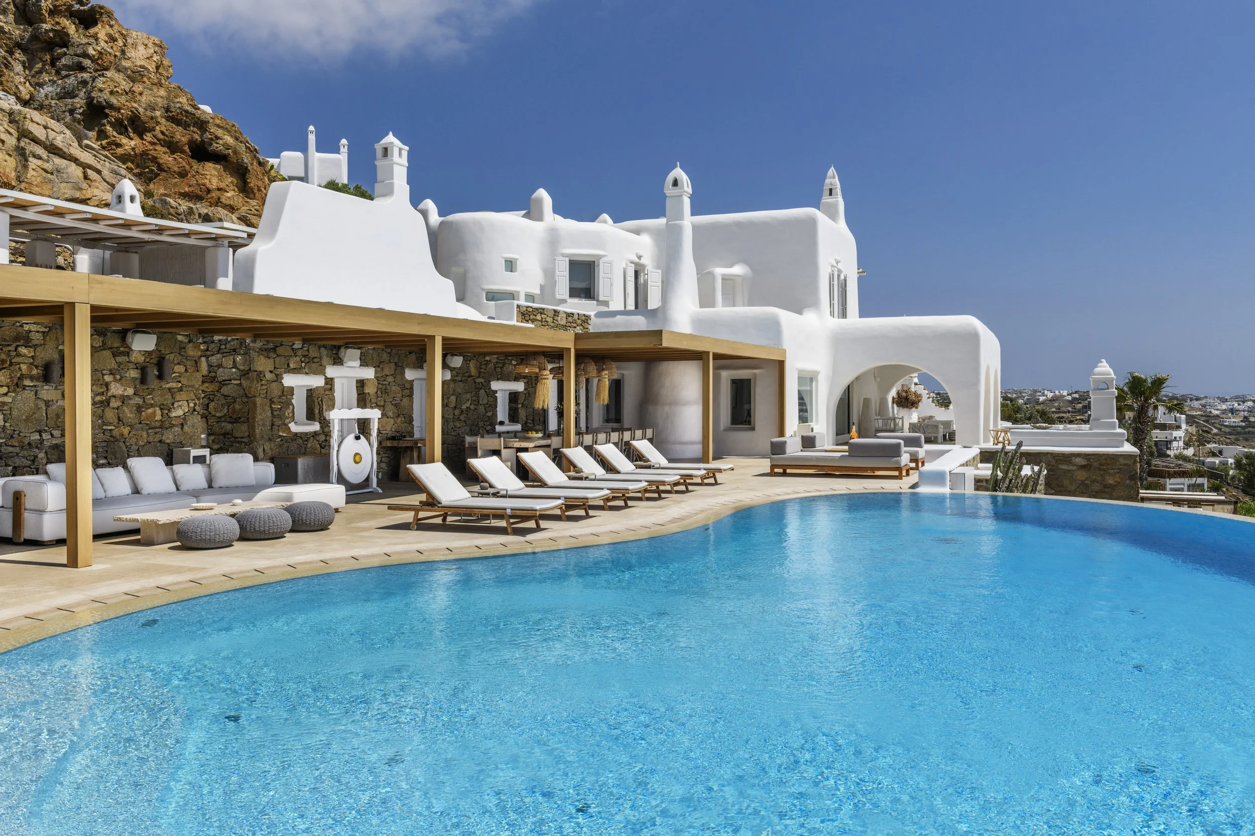 Luxury villa on Mykonos island featuring a large, curved infinity swimming pool, a covered outdoor lounge area with sun loungers, and traditional white Cycladic architecture