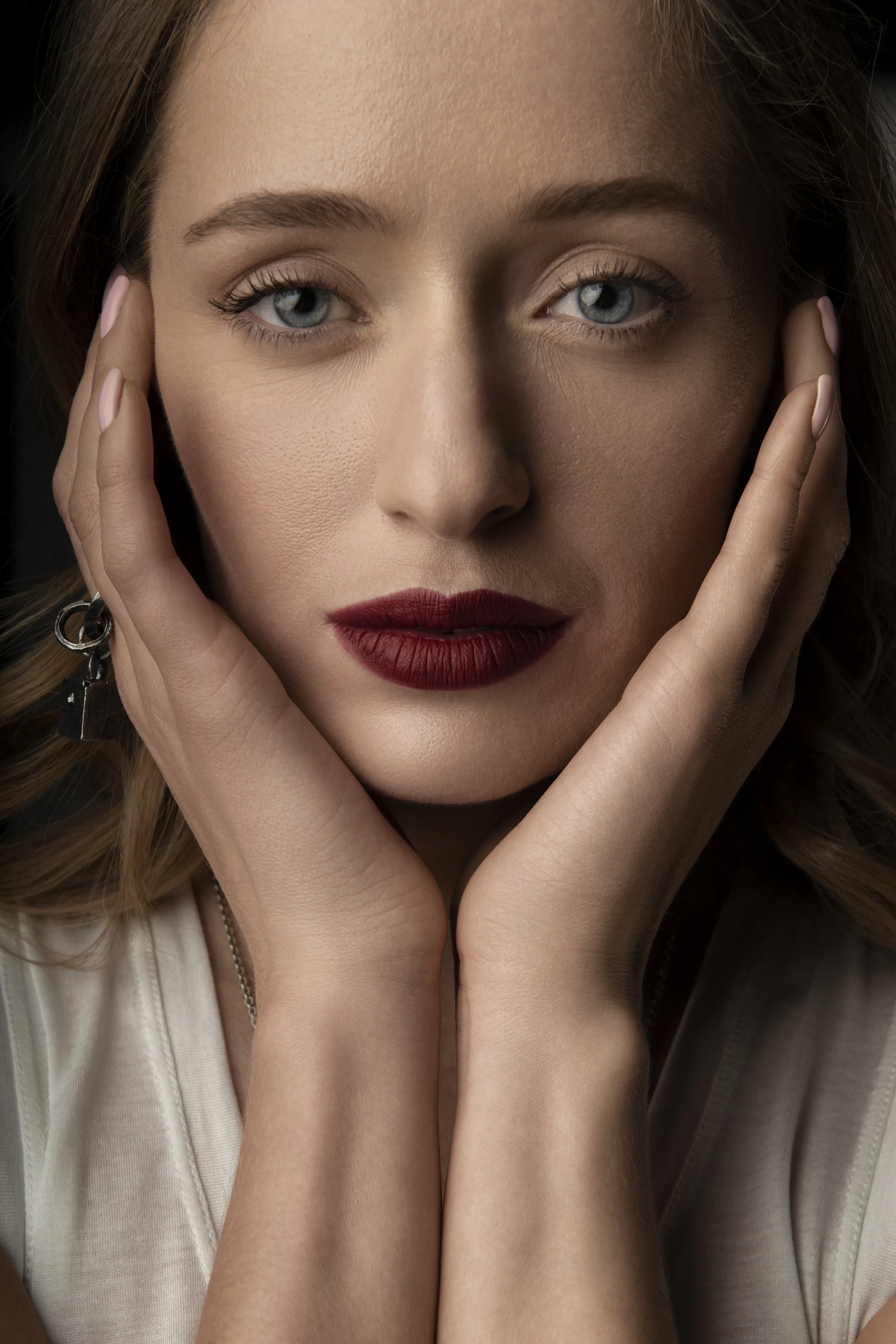 Close-up of a woman with blonde hair and blue eyes, touching her face with both hands, wearing makeup with red lipstick, and a white top.