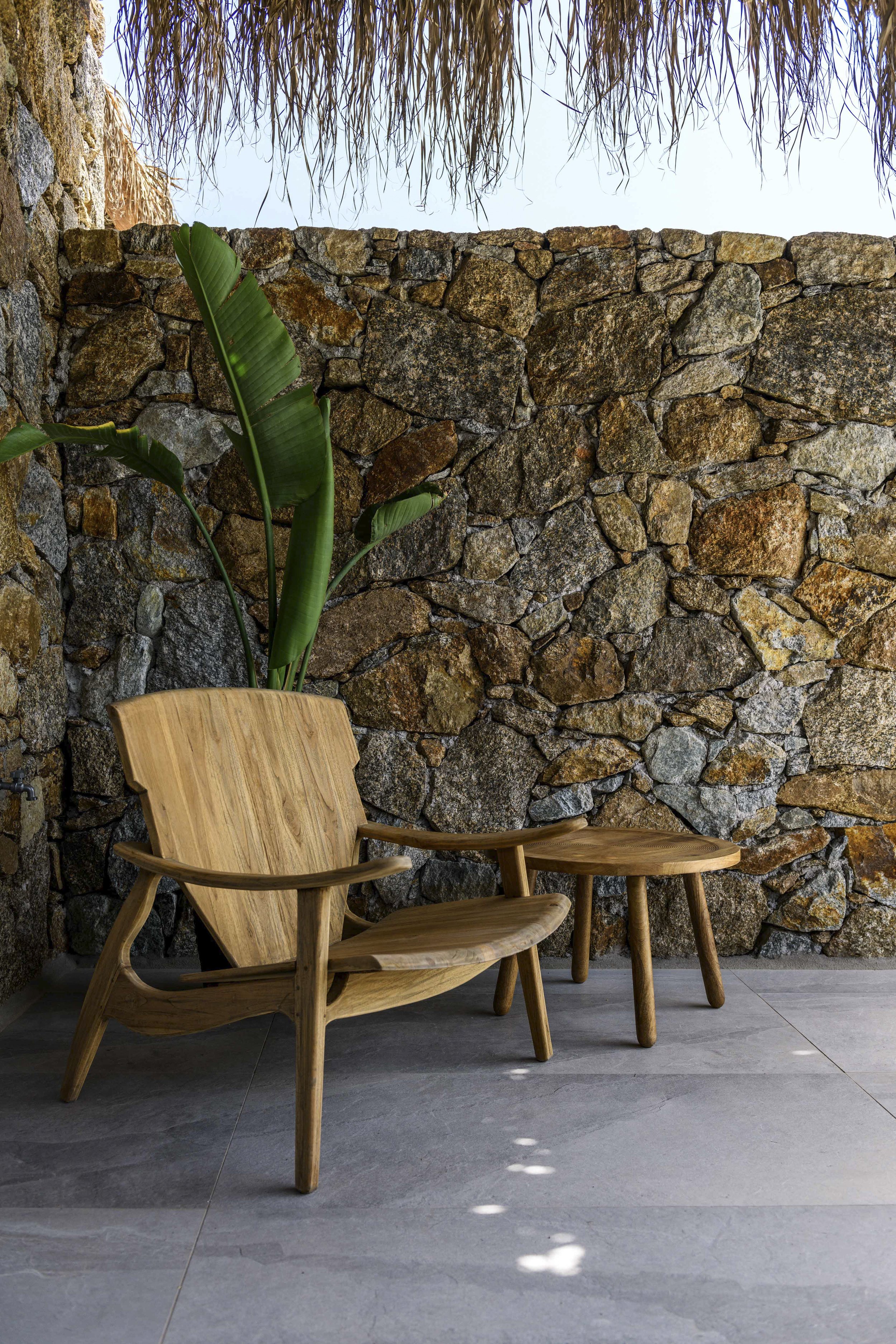 Indoor setting with a stone wall, a tall green plant, and two wooden chairs on a gray tiled floor.