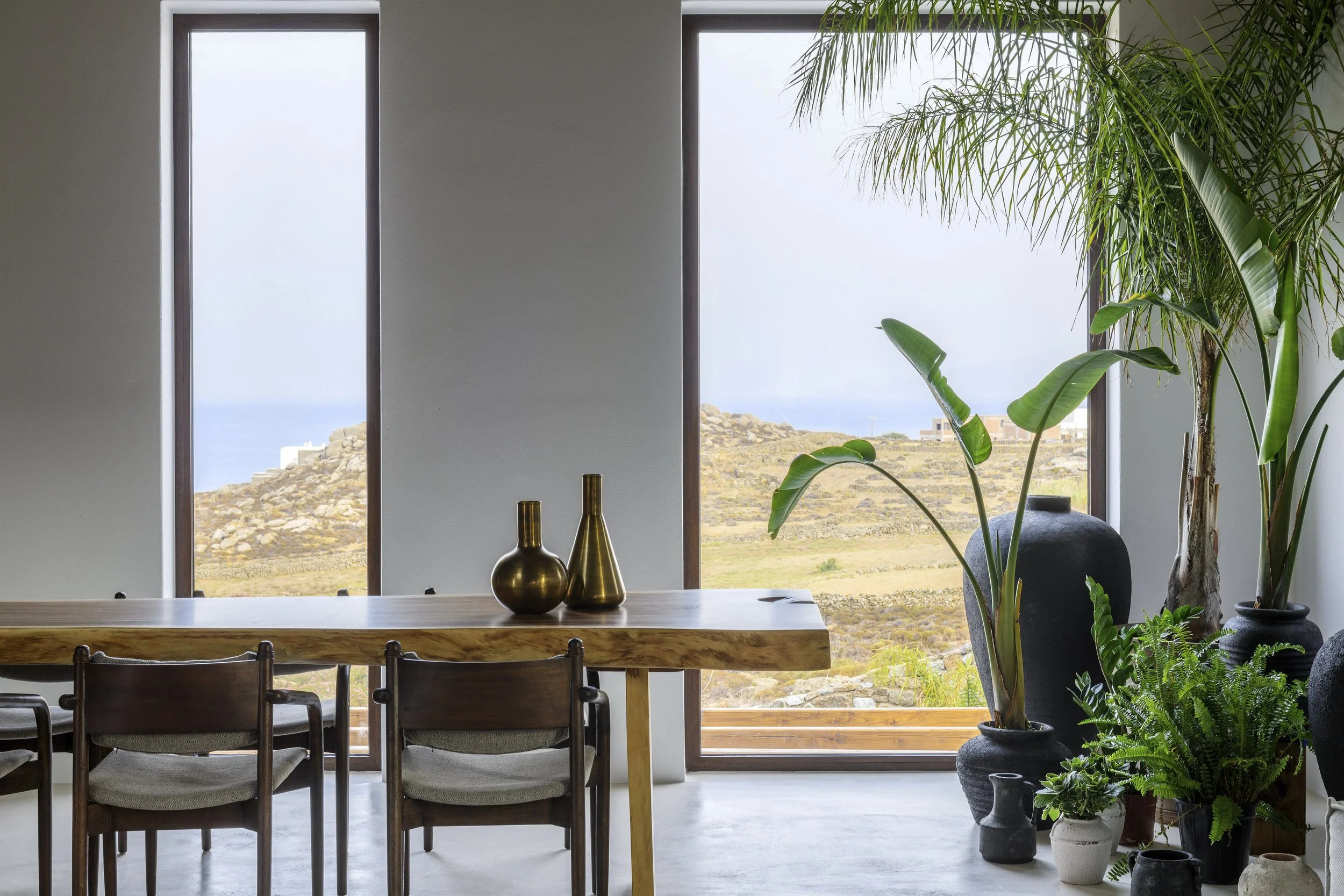 A modern dining area with a wooden table, four chairs, and decorative vases, large windows overlooking a rocky landscape and the ocean, and green plants inside.