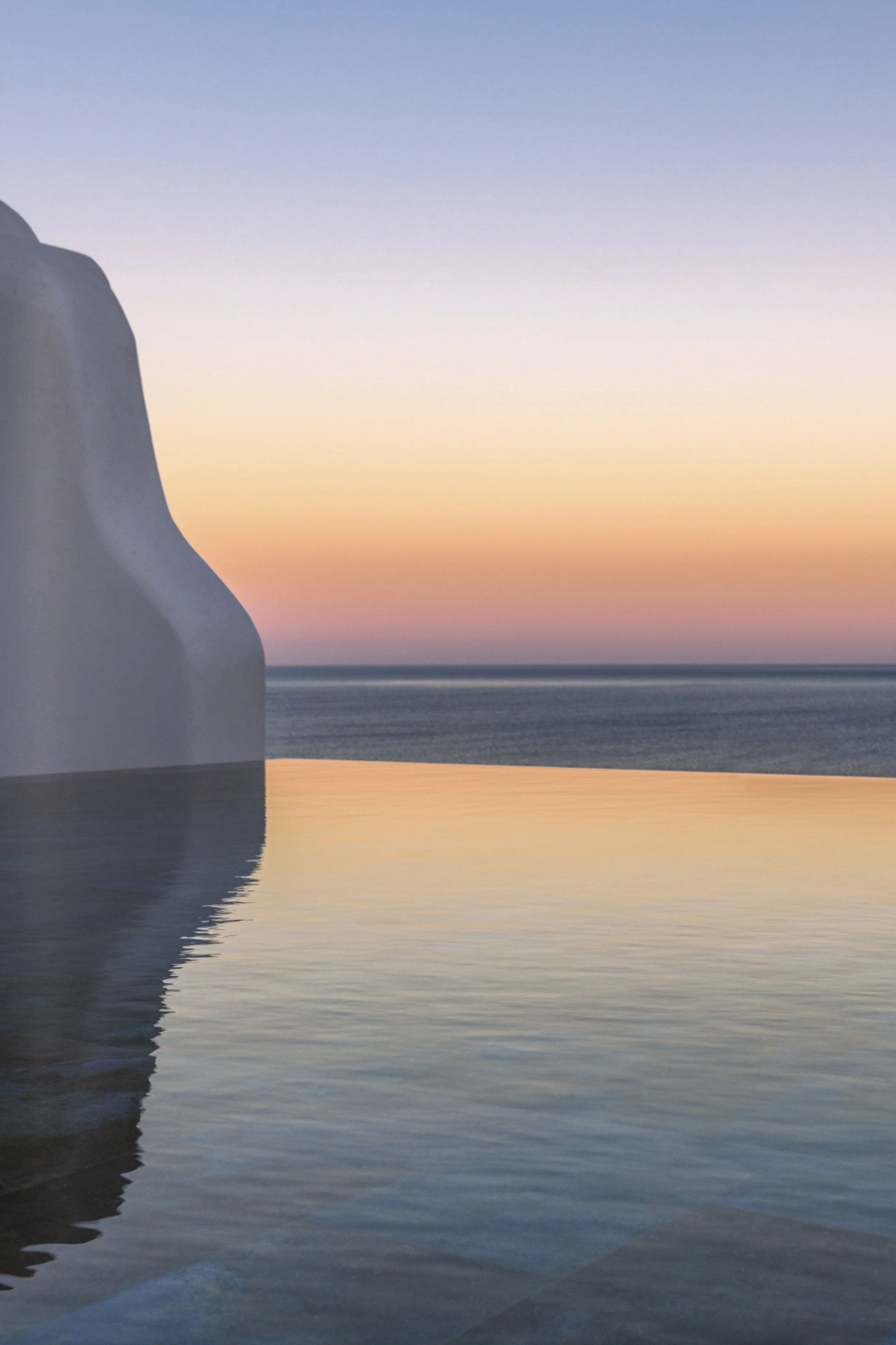 View of white building with curved roof on the left side, reflecting in calm water at sunset with a colorful sky of blue, pink, and orange hues.