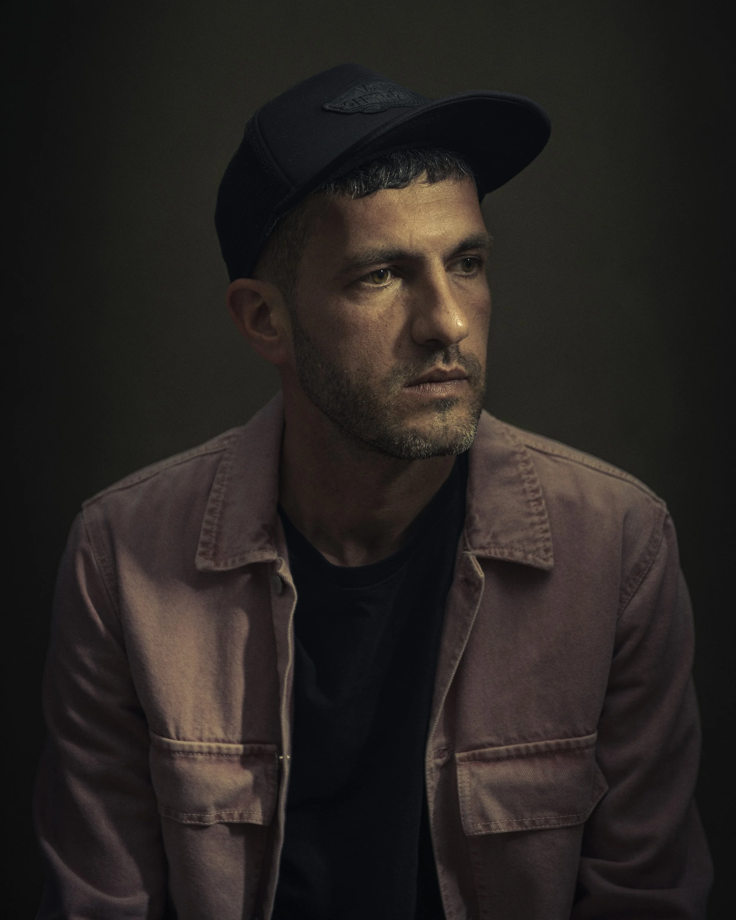 A young man with short dark hair and a beard, wearing a black baseball cap and a pinkish-brown jacket, looking slightly to the right against a dark background.