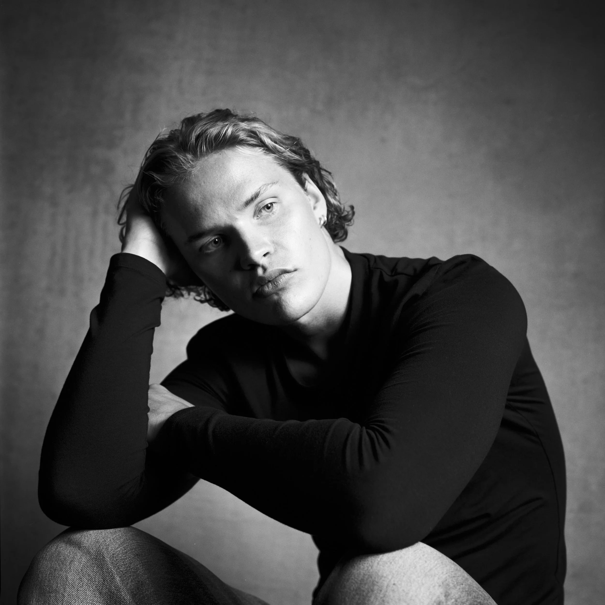 A black-and-white photo of a young man with curly hair resting his head on his hand, wearing a dark long-sleeve shirt, with a neutral expression, against a plain background.