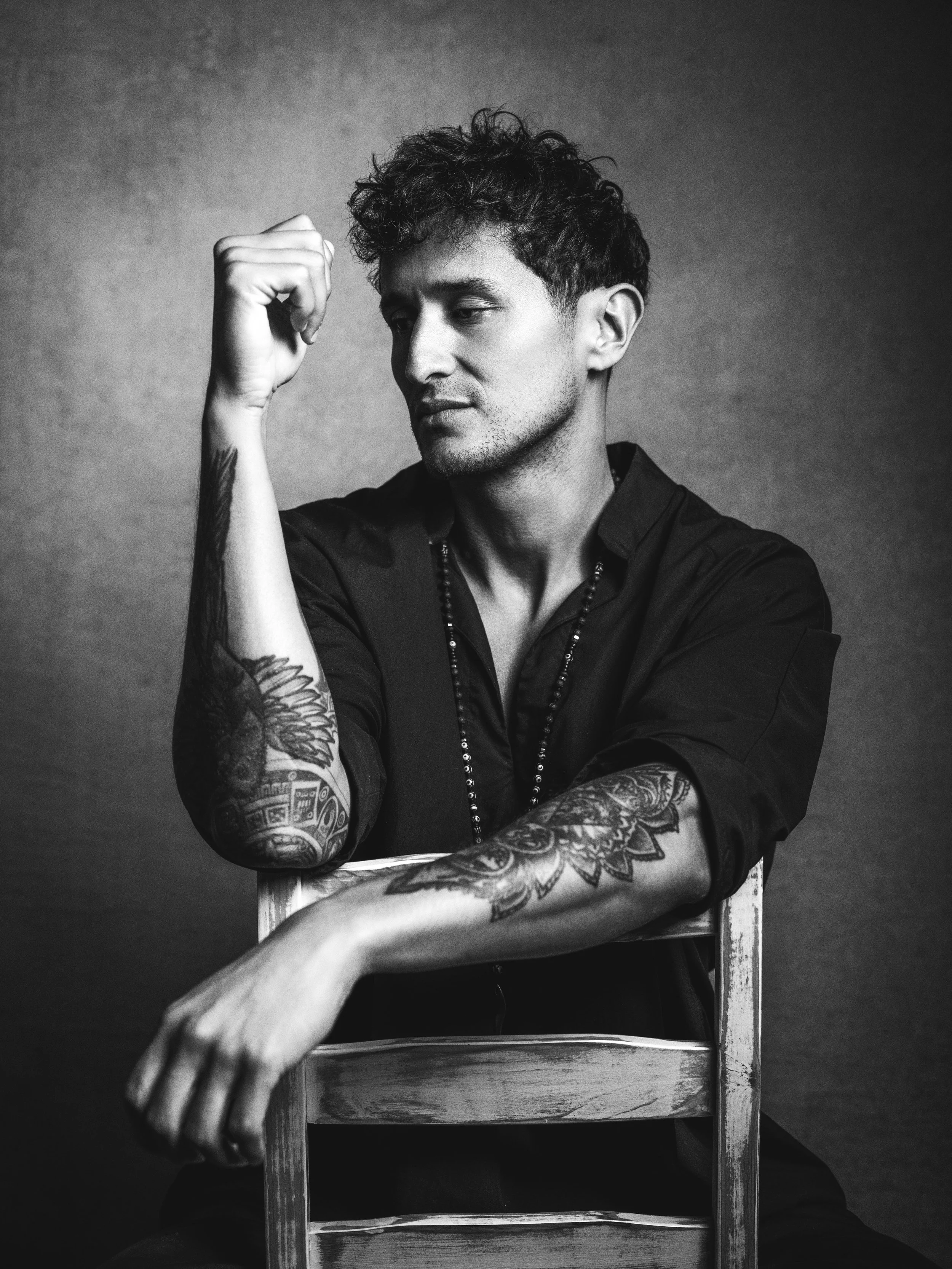 Black and white photo of a man with tattoos on his arm, sitting behind a wooden chair or table, looking down thoughtfully, with his fist raised near his head.