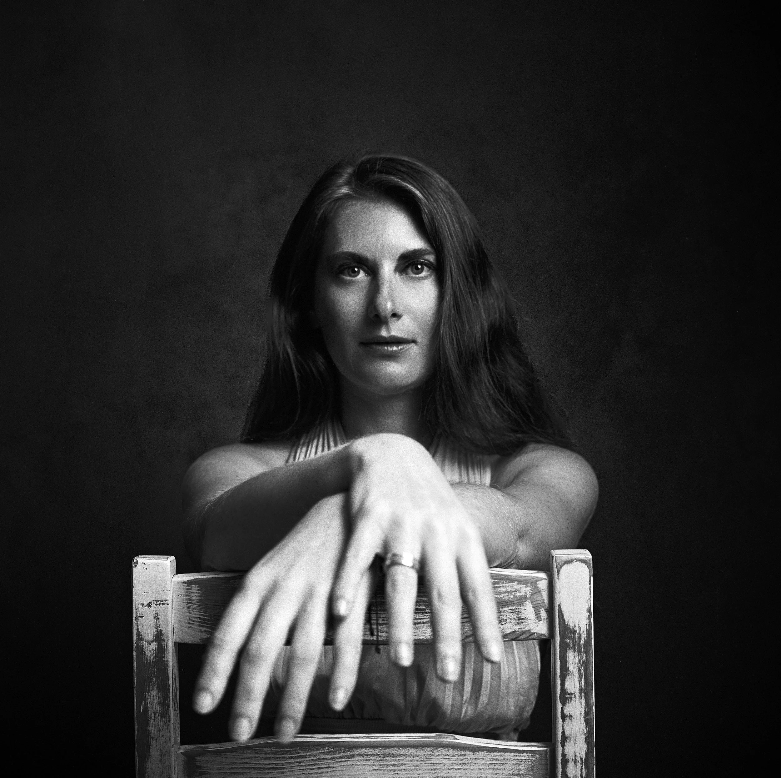 Black and white photograph of a woman with long hair, sitting on a wooden chair with her arm extended forward, looking directly at the camera.