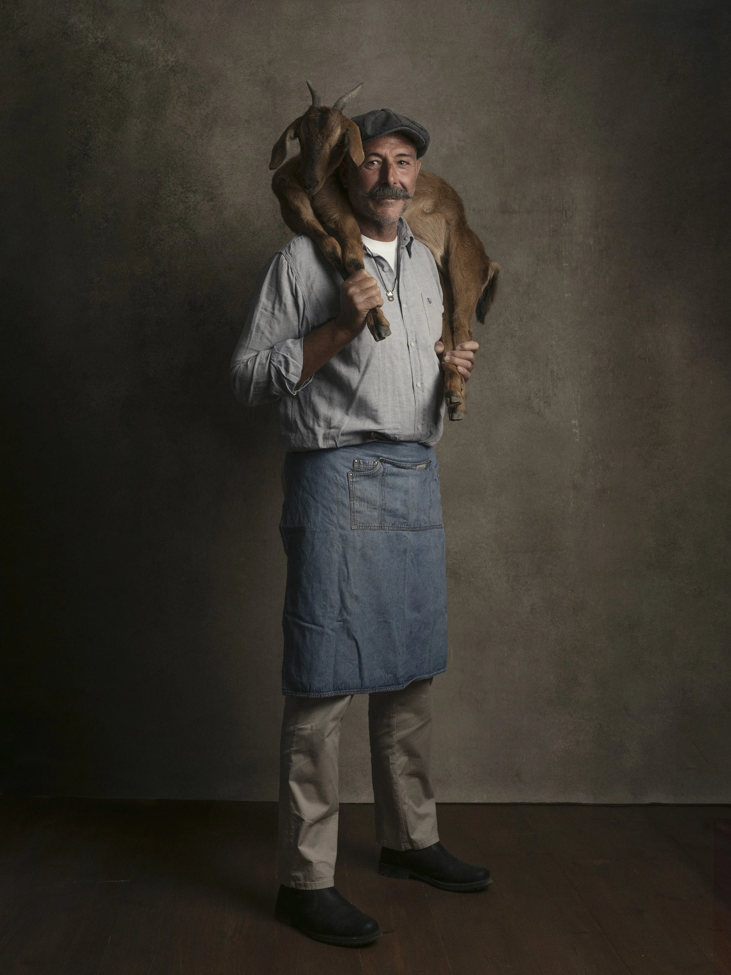 A man wearing a gray shirt, denim apron, and gray cap carrying a brown goat on his shoulder