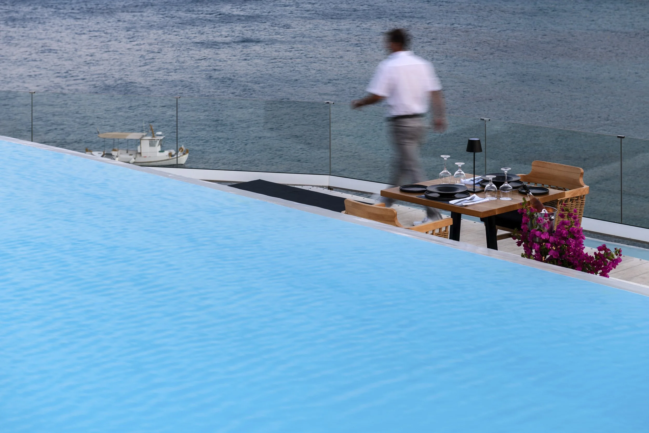 A man in a white shirt walking on a deck overlooking the water with a small boat floating nearby. There is a table set with black plates and glasses, and a vase with pink flowers.