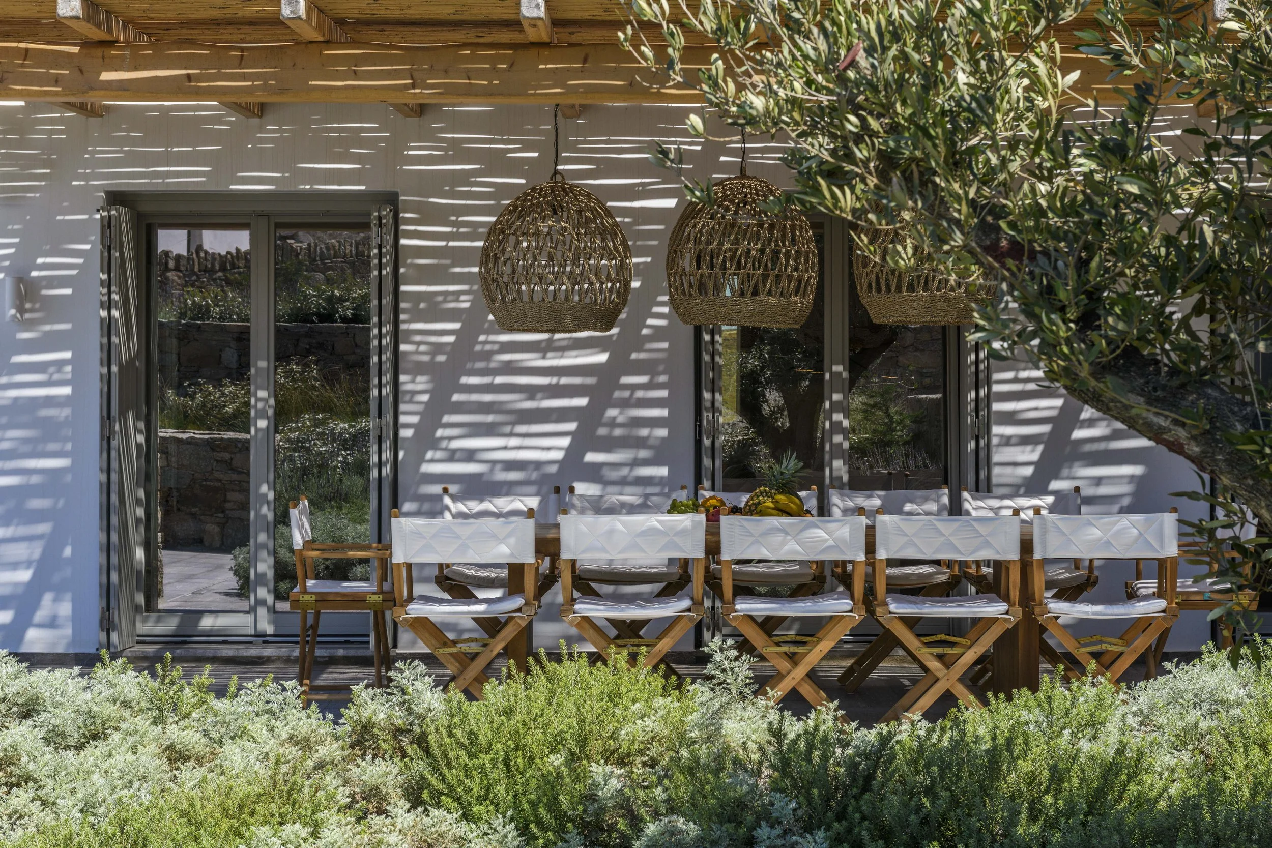 An outdoor dining area with a long table and white chairs, shaded by a pergola with hanging wicker pendant lights. A tree with green leaves is on the right side, and a garden with greenery is in the foreground.