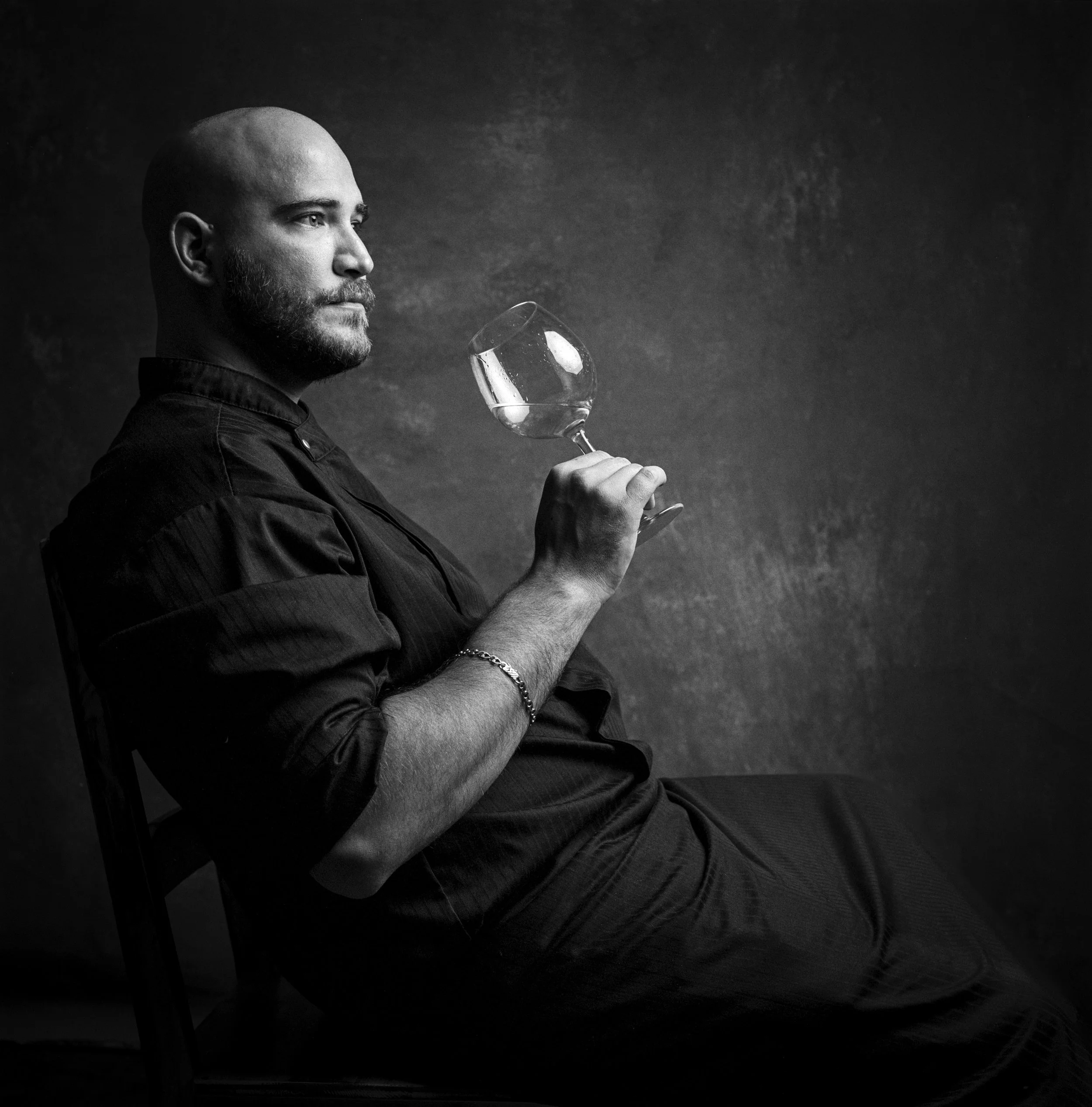 Black and white photo of a man with a tattooed arm, bald head, and beard, holding a wine glass and looking to the left.