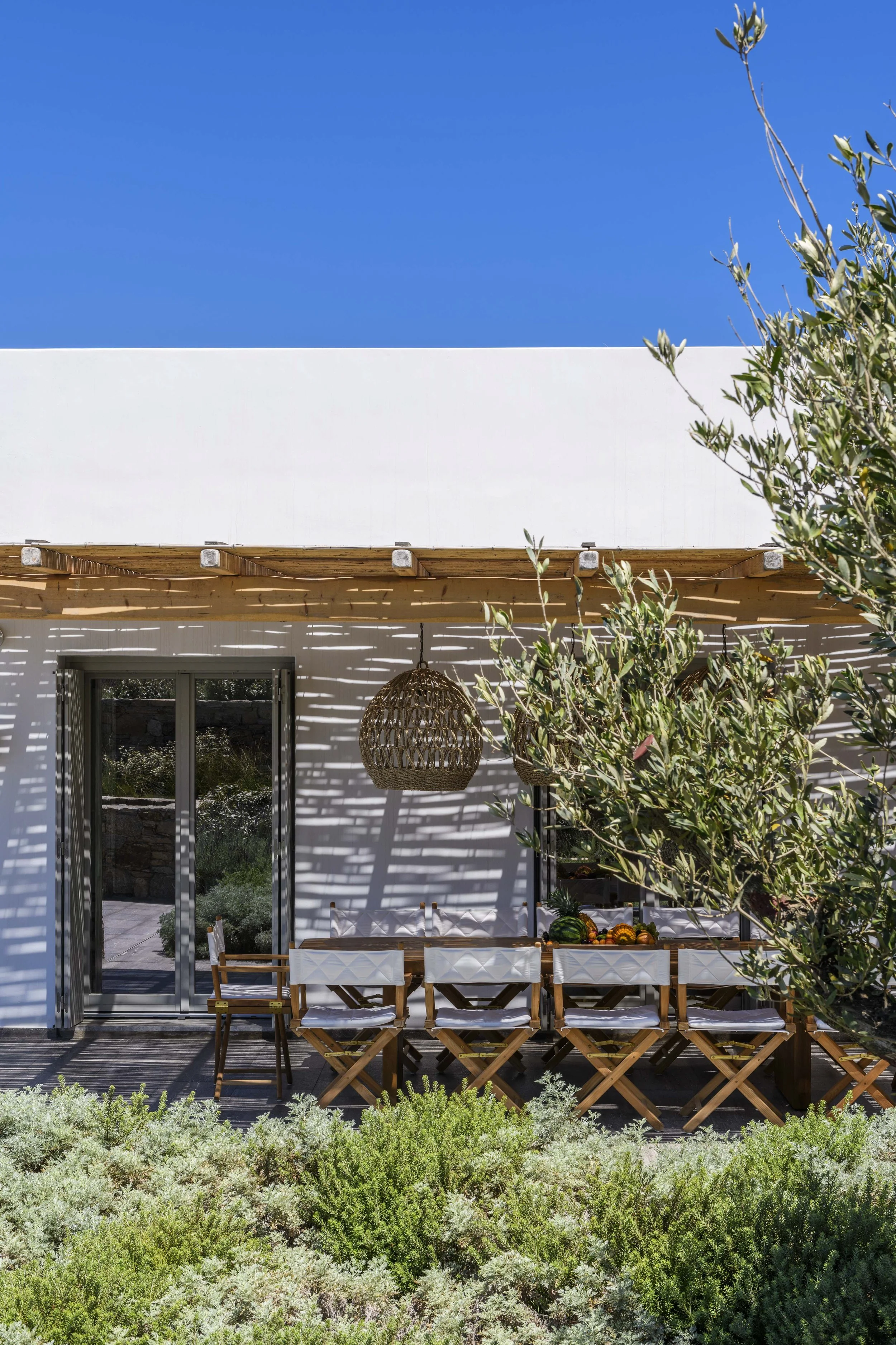 Outdoor patio with a dining table and chairs, hanging wicker lamps, a white wall with a wooden pergola, and greenery nearby.