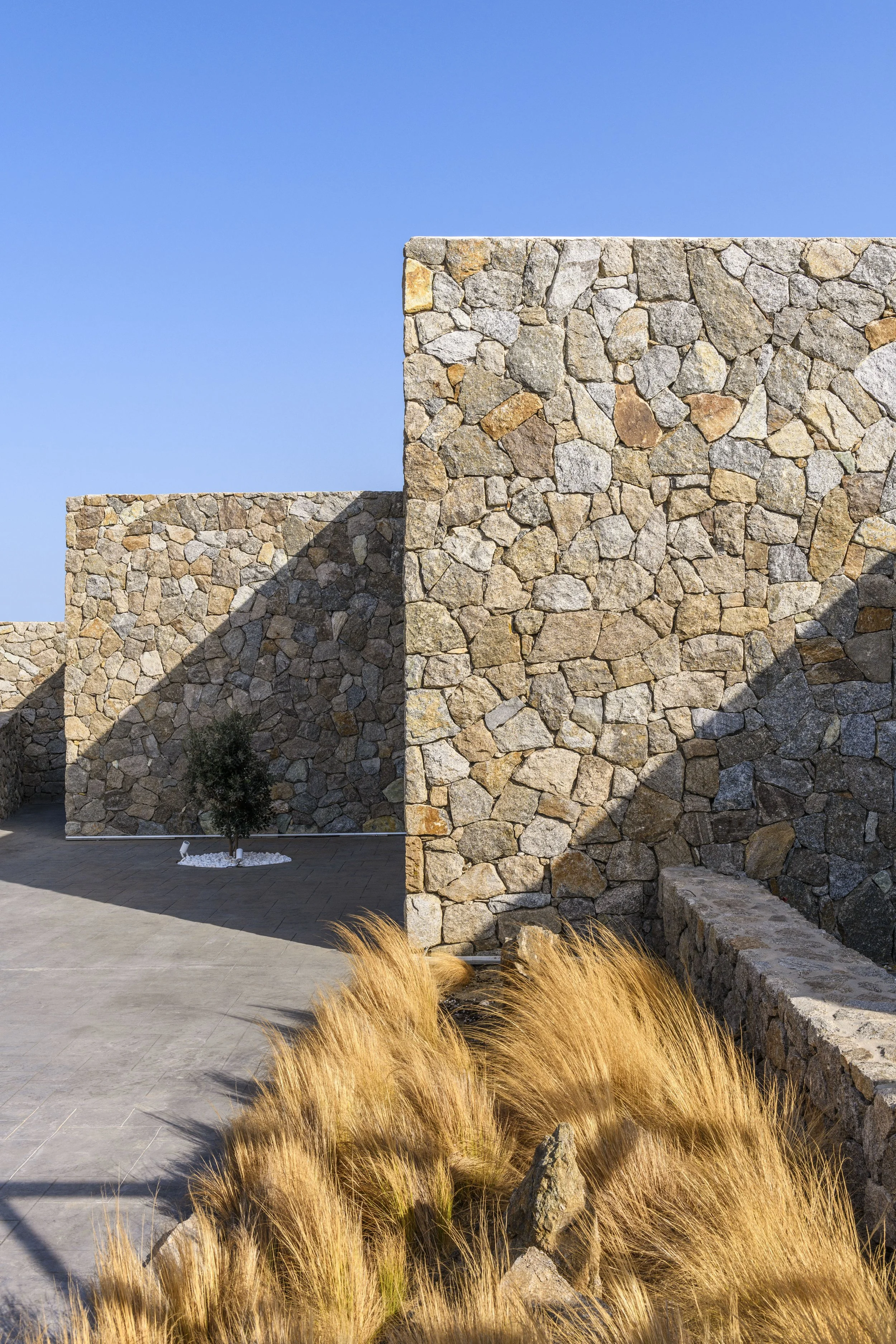 Minimalist luxury villa on Mykonos island, built into a dry Mediterranean hillside with sustainable landscaping, natural stone walls, and large glass windows.