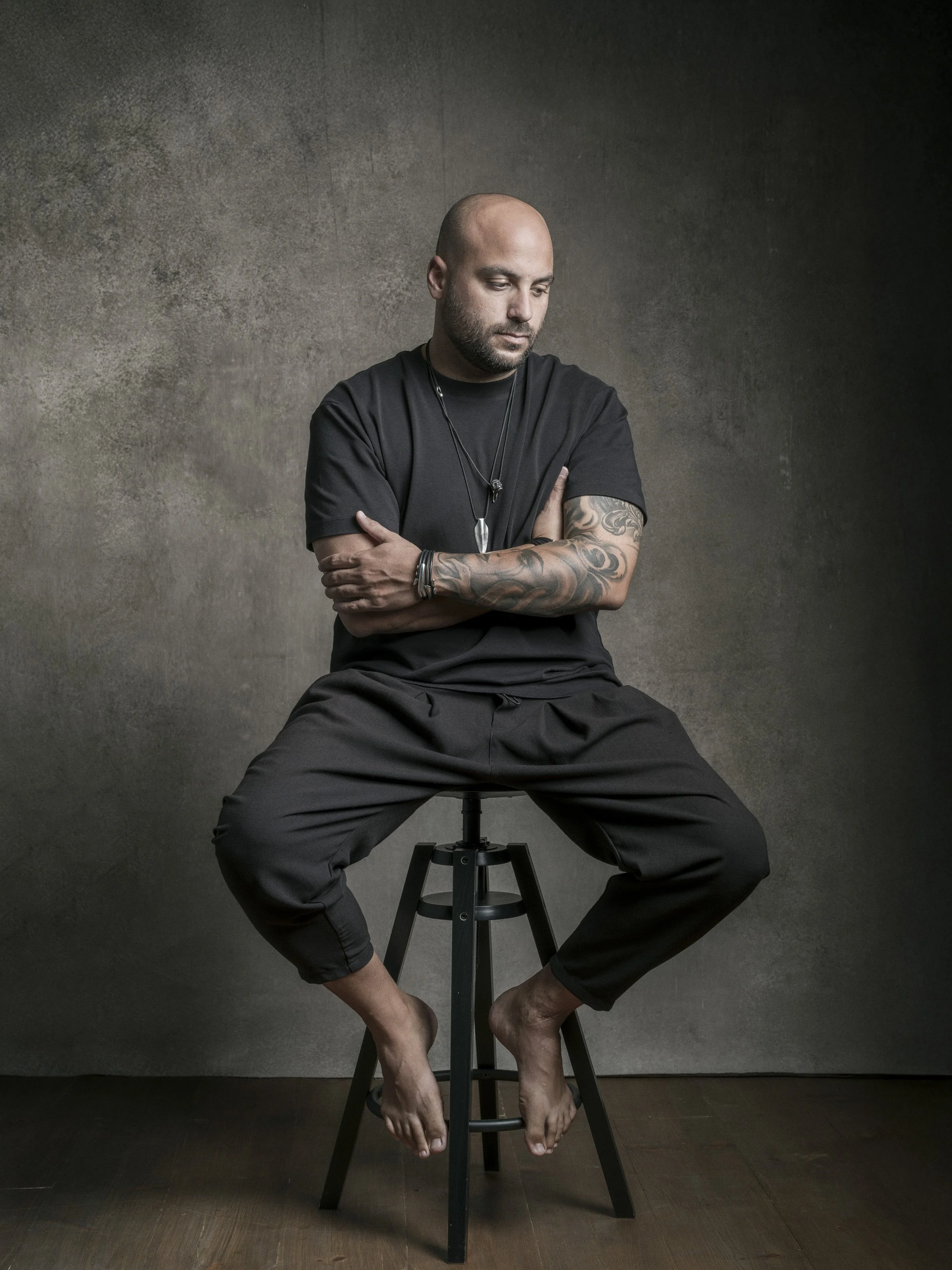 A man with a shaved head and tattoos on his arms, wearing a black shirt and pants, sitting on a stool with his arms crossed, looking down. His feet are bare and his toes are touching the wooden floor. The background is a dark textured wall.