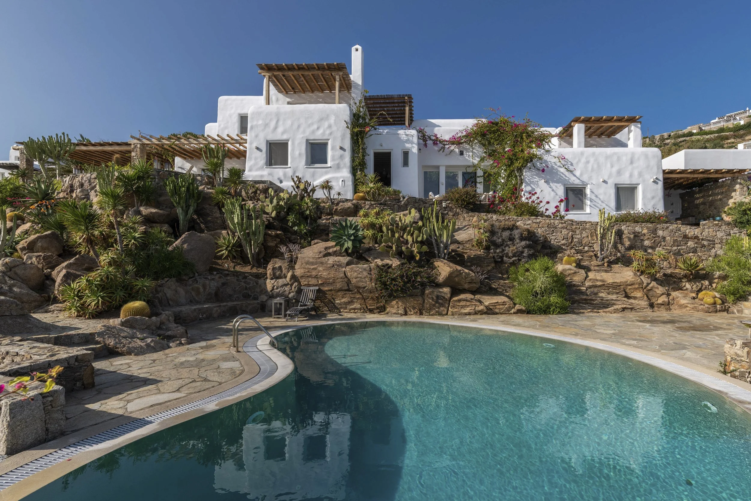 A luxurious house with white walls and wooden pergolas on a hillside, featuring a swimming pool surrounded by rocks and desert plants under a clear blue sky.