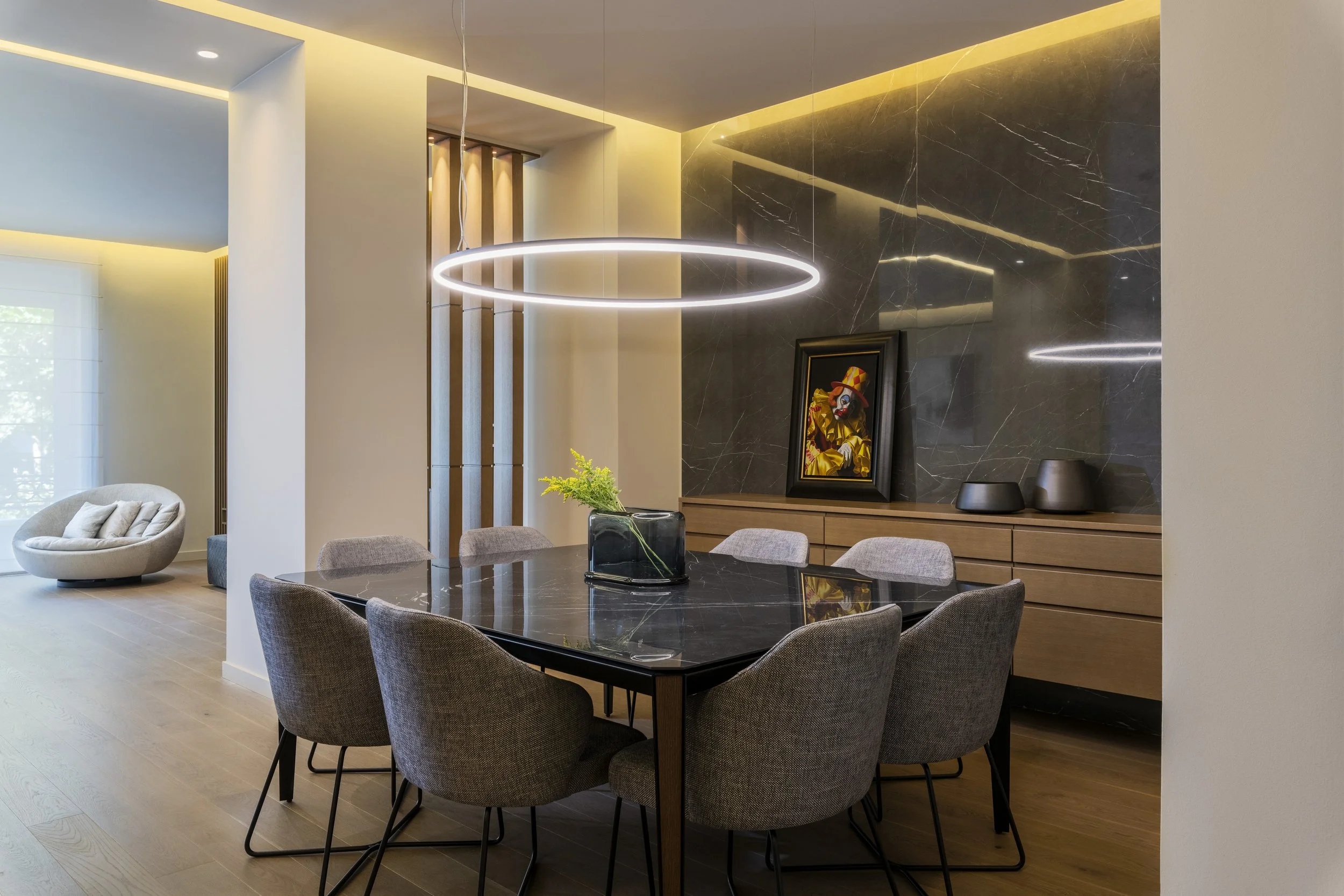 Modern dining room with a black marble table surrounded by gray upholstered chairs, a circular LED hanging light, a black framed clown portrait, and a dark marble accent wall.