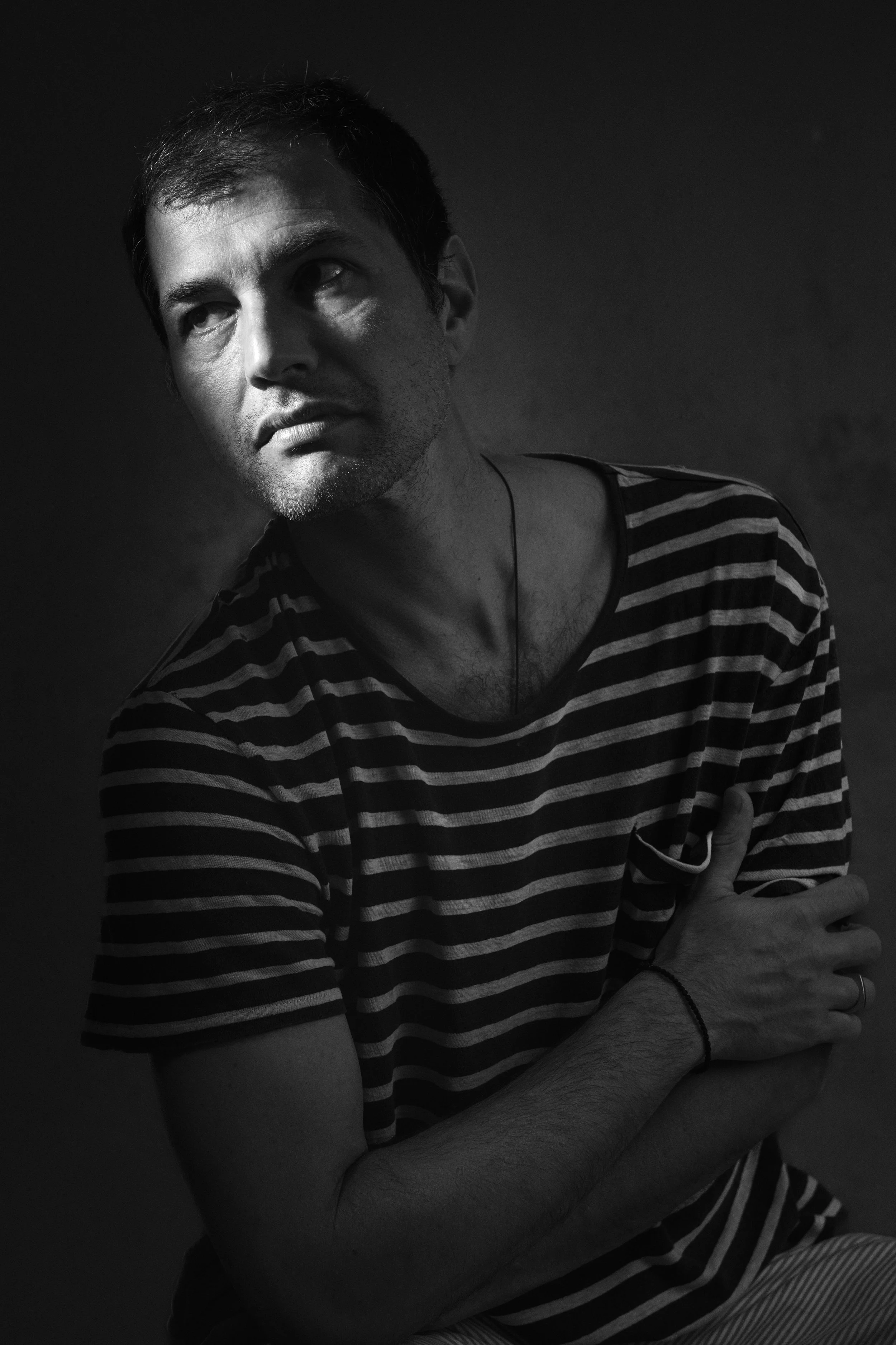 Black and white photograph of a man with short dark hair, wearing a striped shirt, looking to his left. His arms are crossed, with his right hand resting on his left arm. The background is dark and plain.