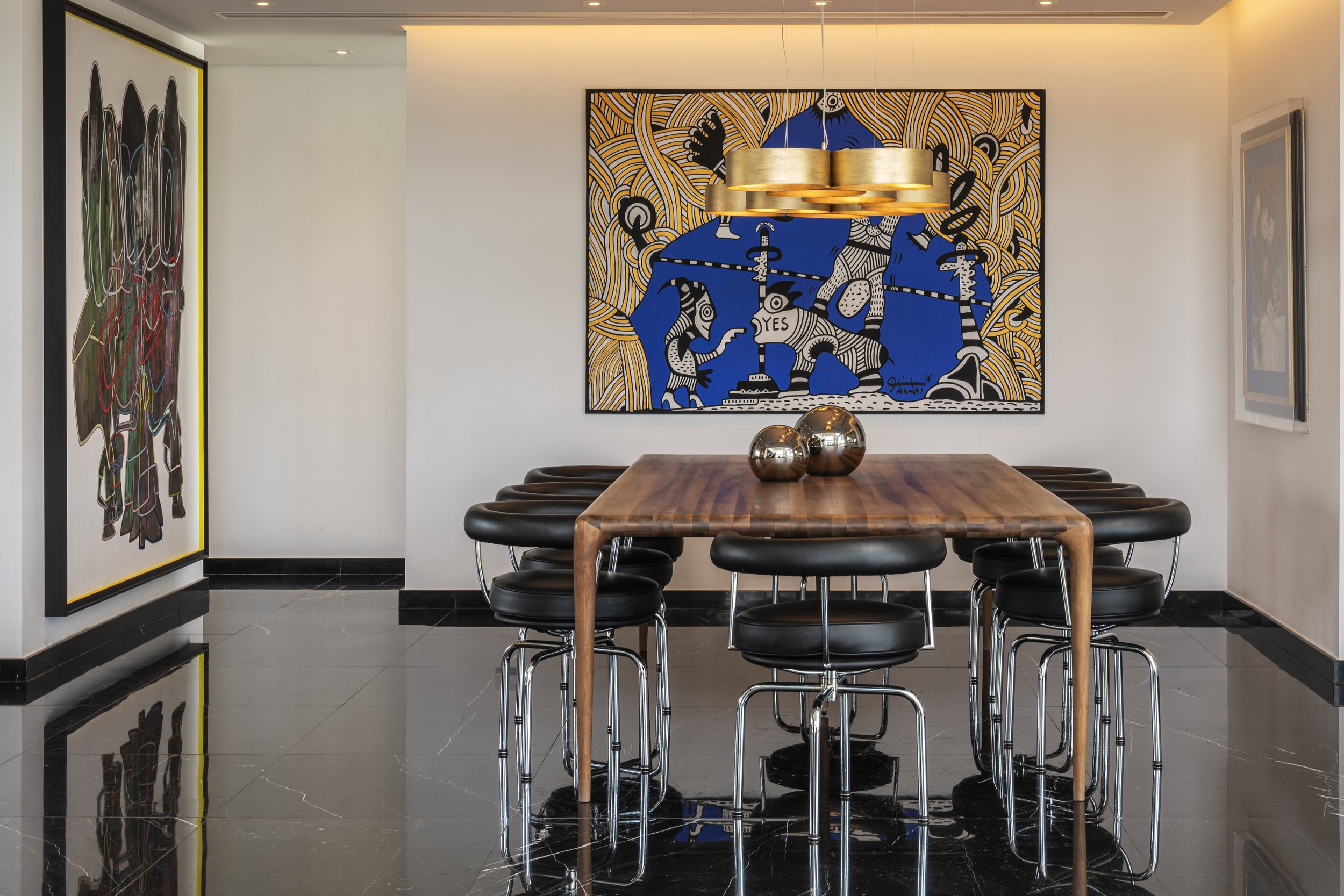 Modern dining room with a wooden table, black chairs, a large colorful abstract wall art, a hanging gold light fixture, and decorative metallic spheres on the table.