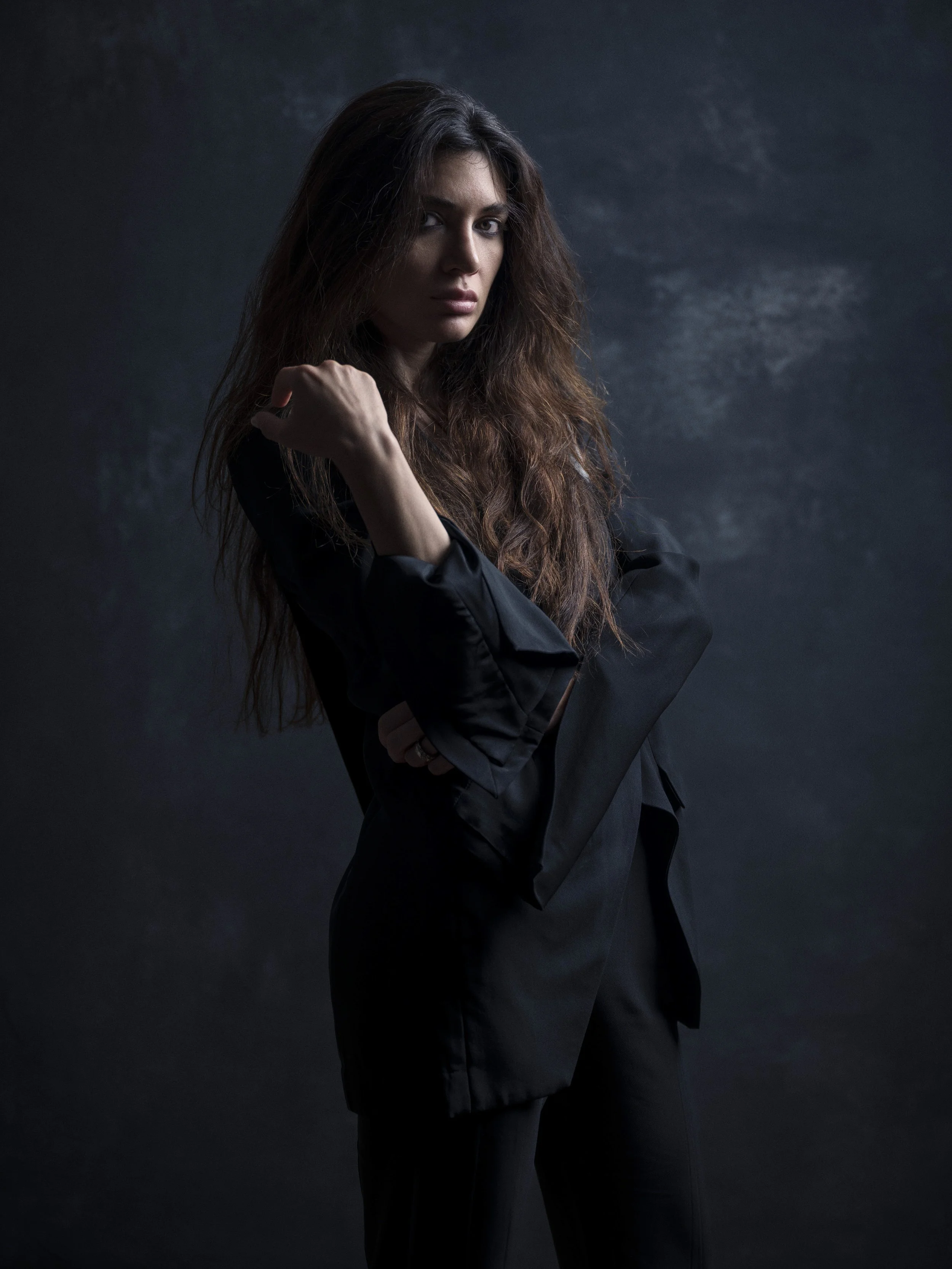 A confident woman with long wavy hair posing against a dark background, wearing a black jacket.