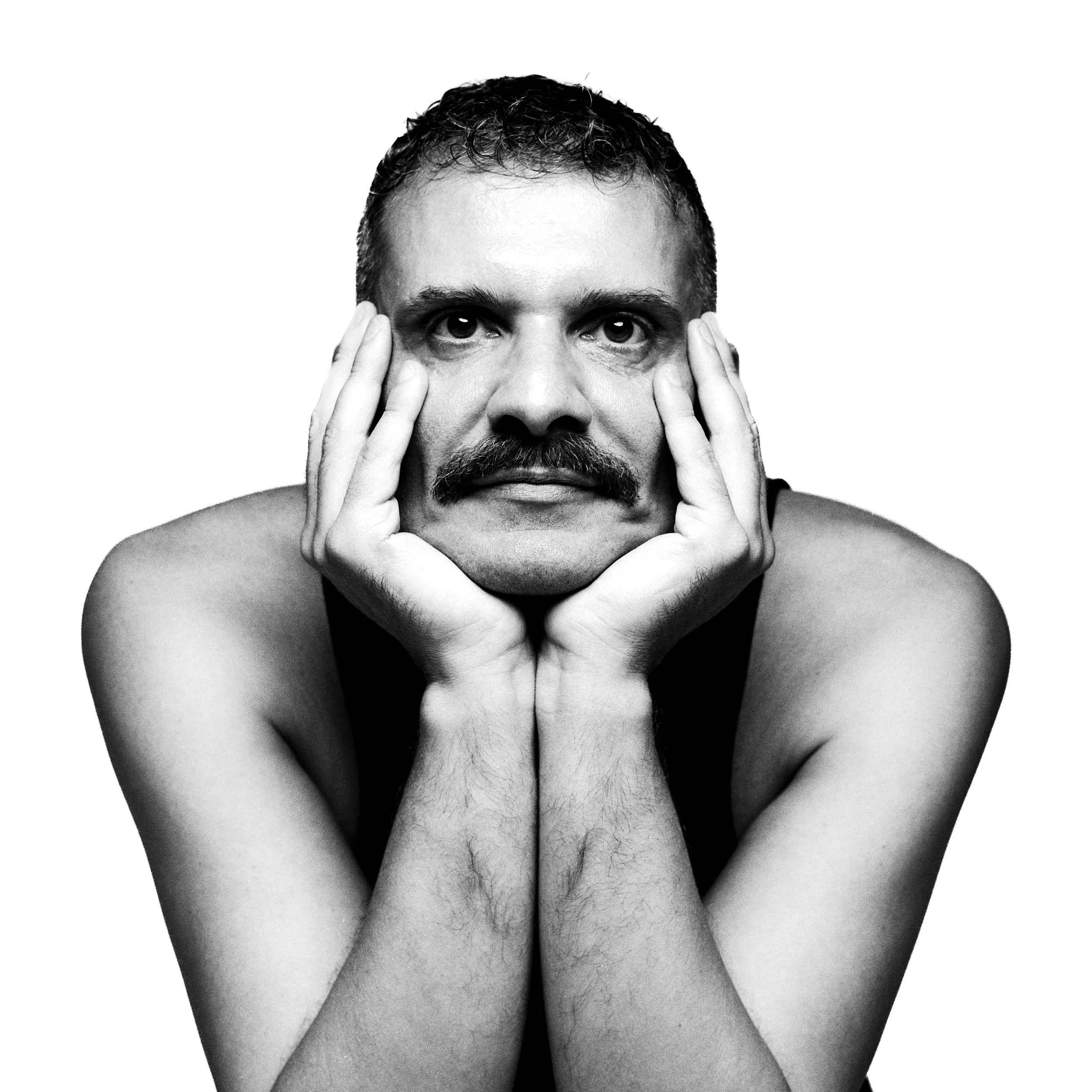 Black and white photo of a man with curly hair and a beard, holding his face with both hands, looking directly at the camera.