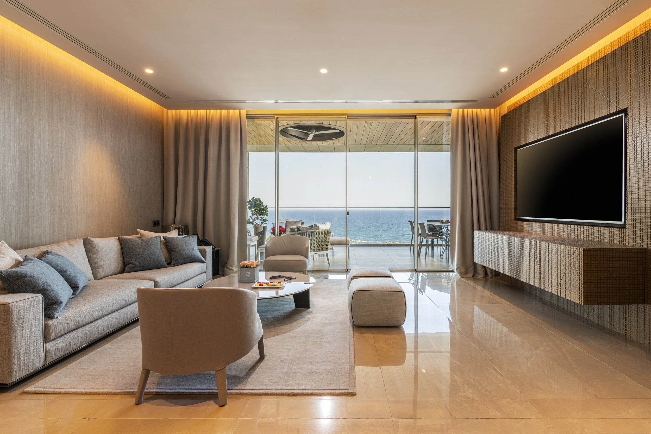 Living room with beige sofa, gray throw pillows, beige armchair, round coffee table with yellow flowers, and large glass doors leading to a balcony with ocean view.
