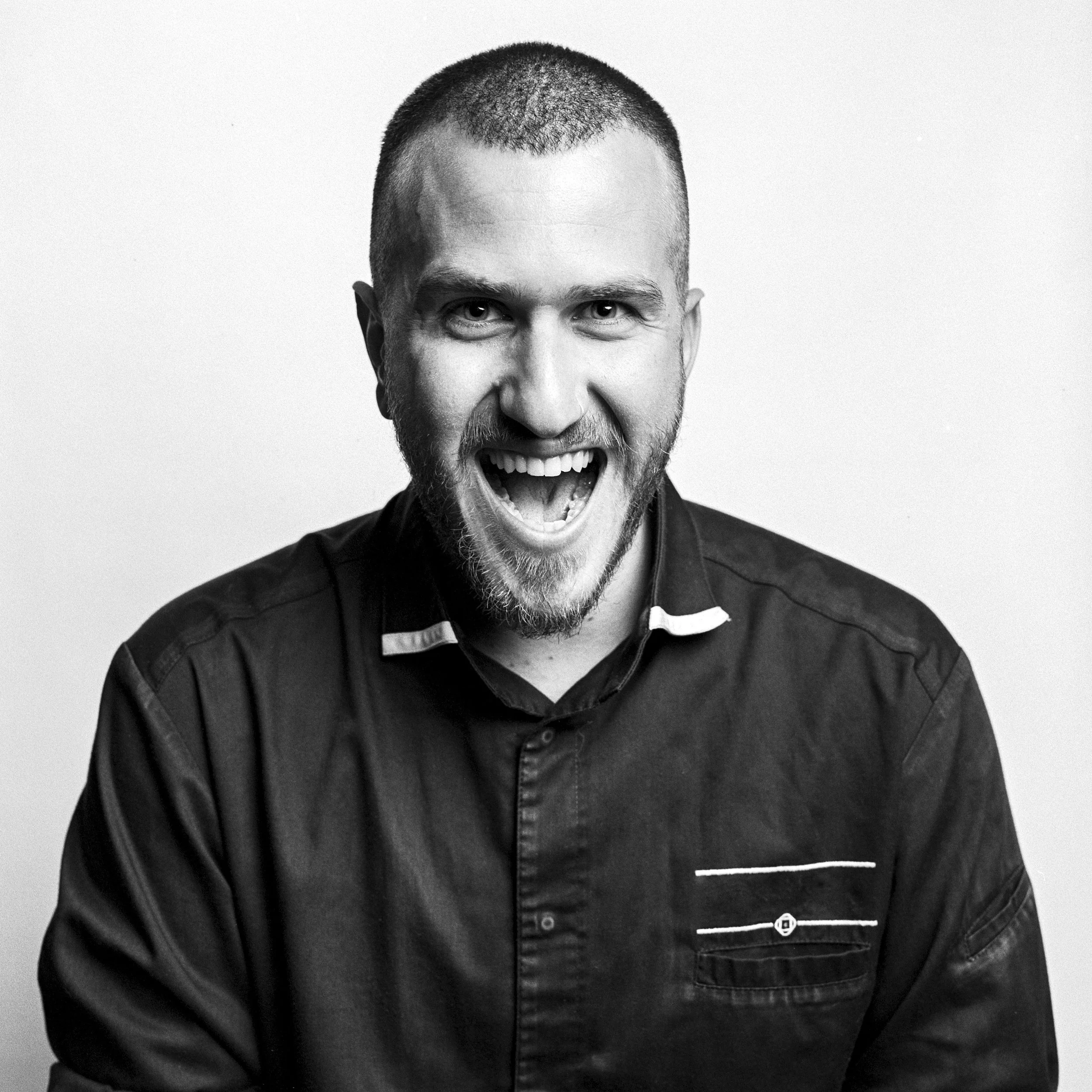 A black and white photo of a man with a beard and short hair, smiling widely and showing his teeth, wearing a dark collared shirt with a striped design.