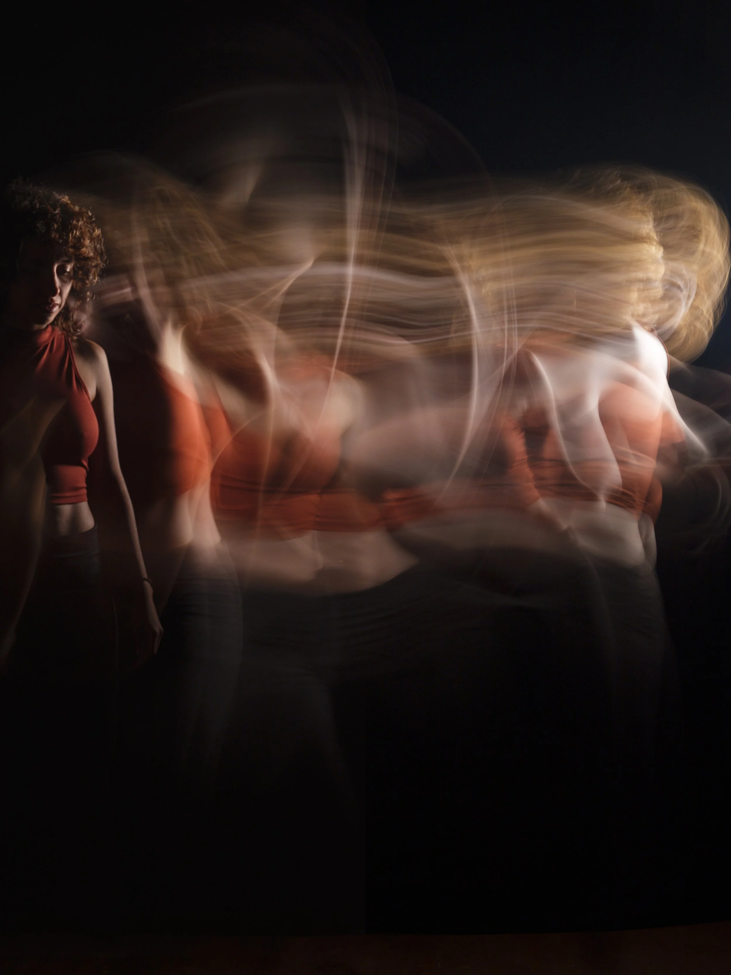 A person with curly hair wearing a sleeveless top, standing against a dark background with long exposure light trails flowing behind them.