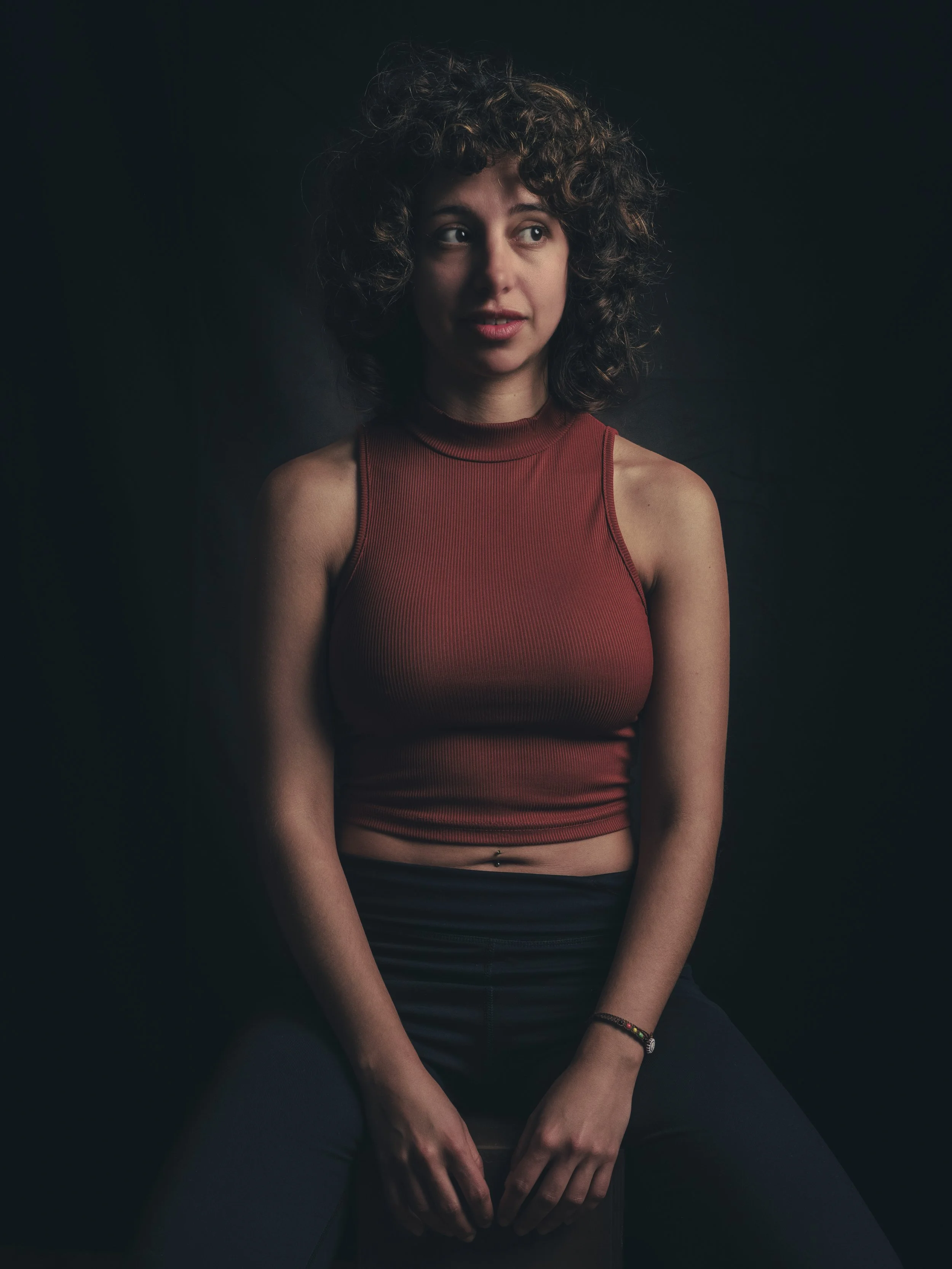 A young woman with curly hair wearing a sleeveless red top and black pants sitting against a dark background, with her hands resting on her lap.