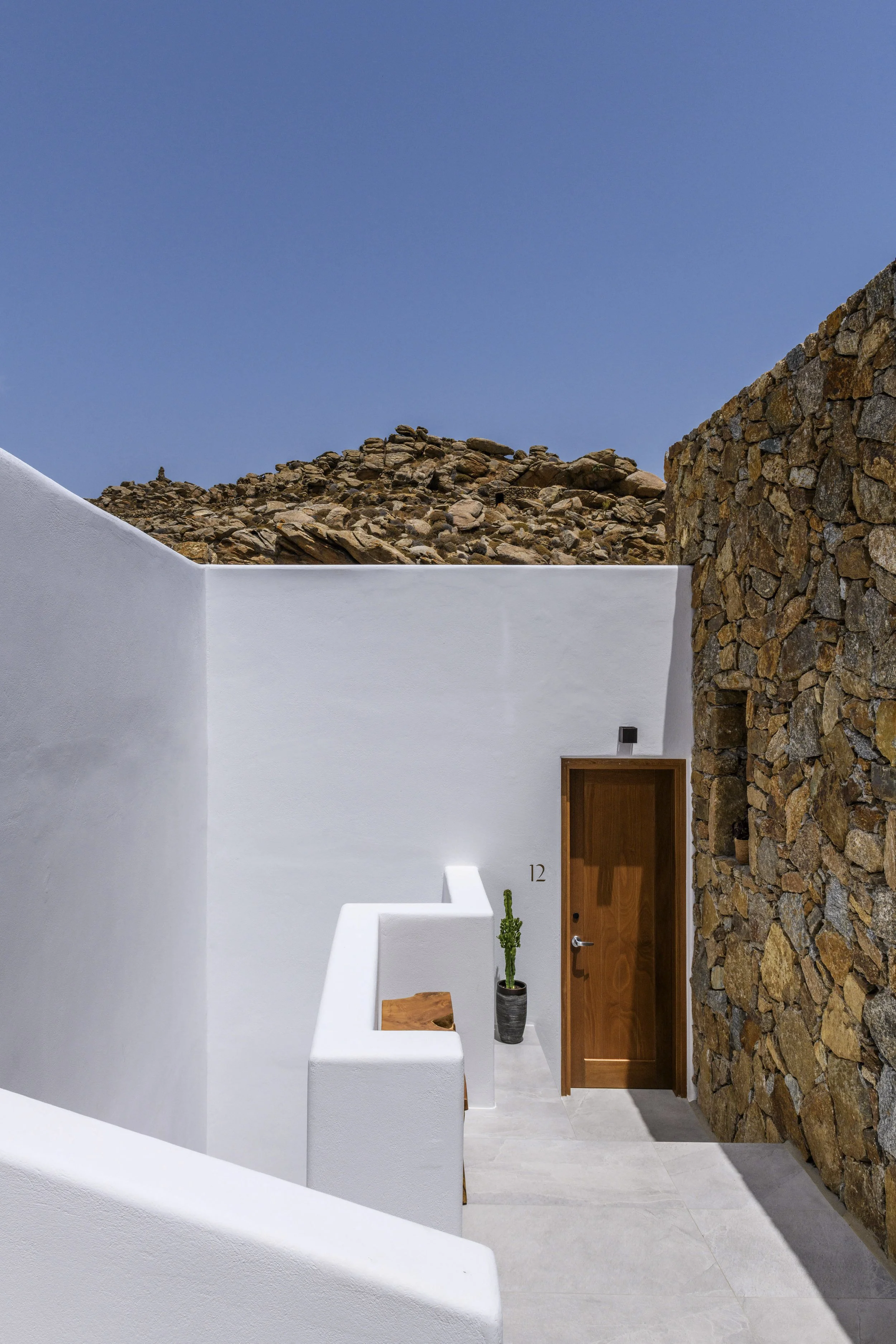 A minimalist outdoor courtyard with white walls, a wooden door, a potted plant, and a rocky hillside in the background under a clear blue sky.