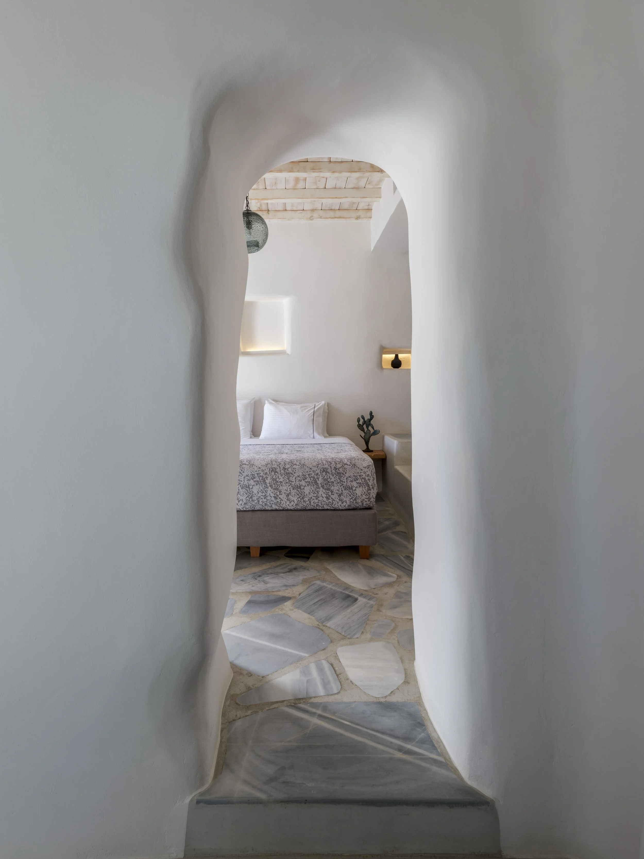 A view through a curved, white plaster door frame into a bedroom with a bed, pillows, and a nightstand with decorative items. The ceiling is wooden with exposed beams, and the floor is made of patterned stone tiles.