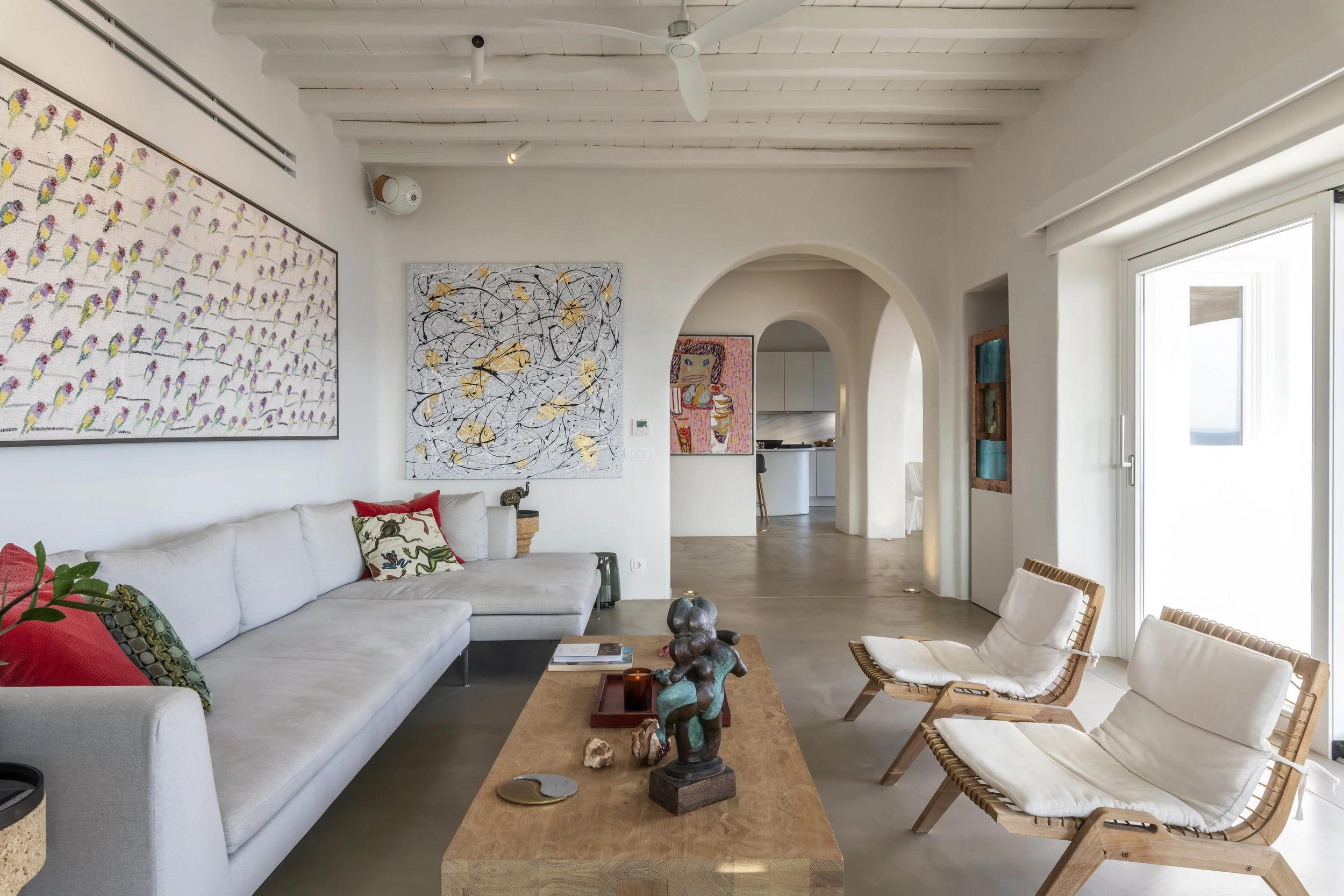 Living room with white sofa, colorful pillows, art on the wall, wooden coffee table with sculptures, and chairs near glass door, bright with natural light.