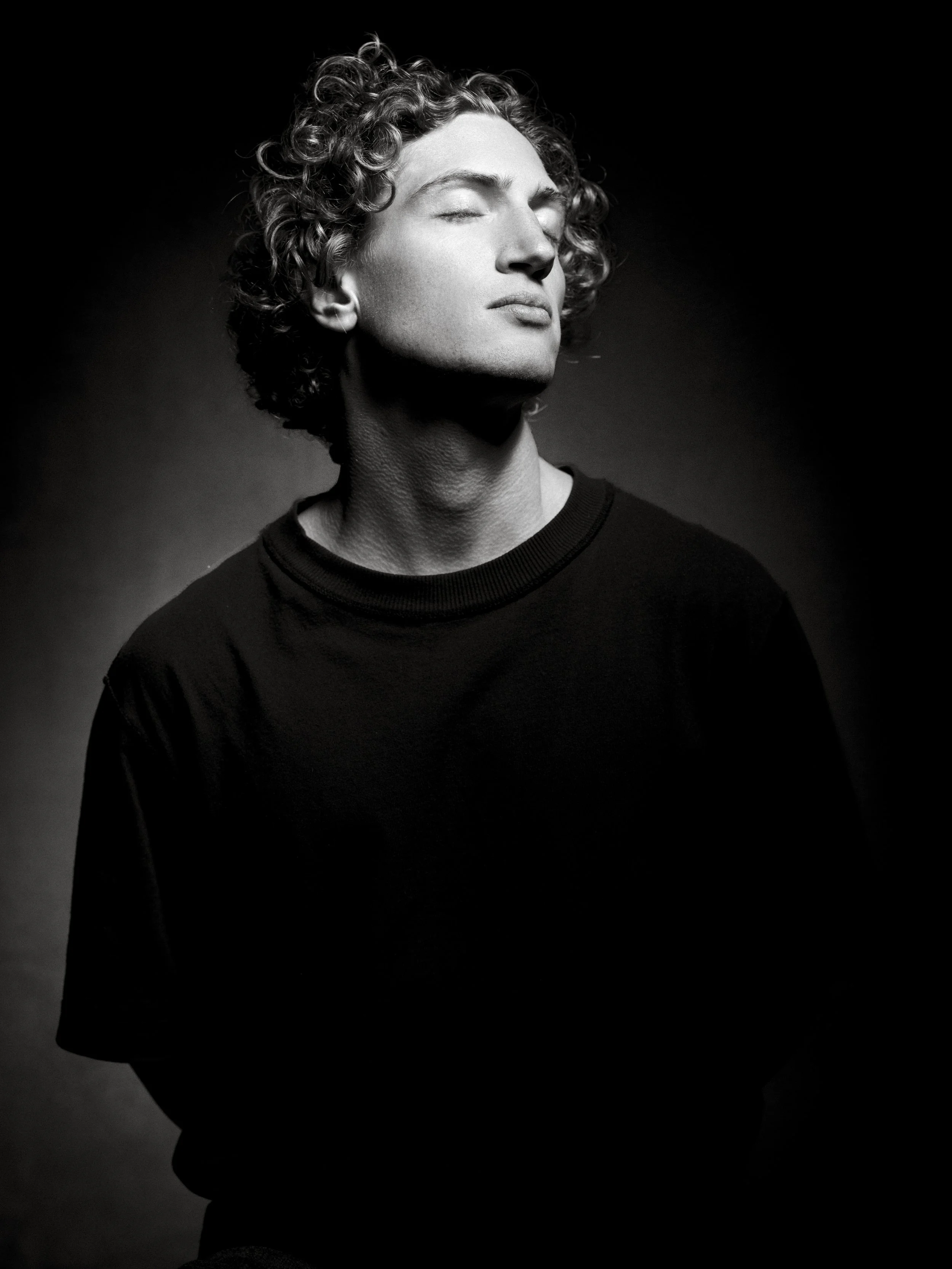Black and white portrait of a person with curly hair, eyes closed, wearing a black shirt, in a dramatic lighting setup.