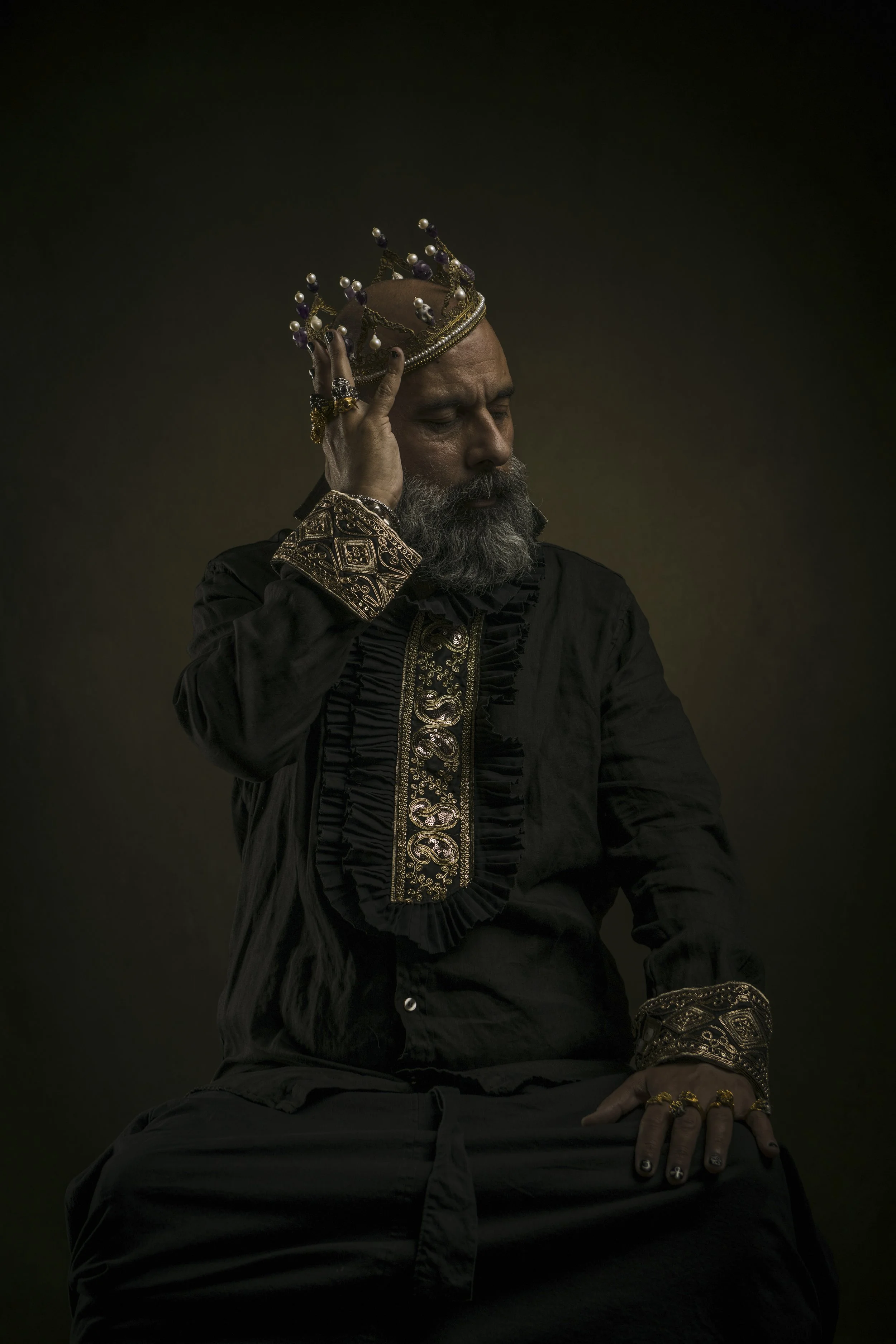 A man with a beard dressed in black, wearing a crown and ornate jewelry, seated with eyes closed against a dark background.