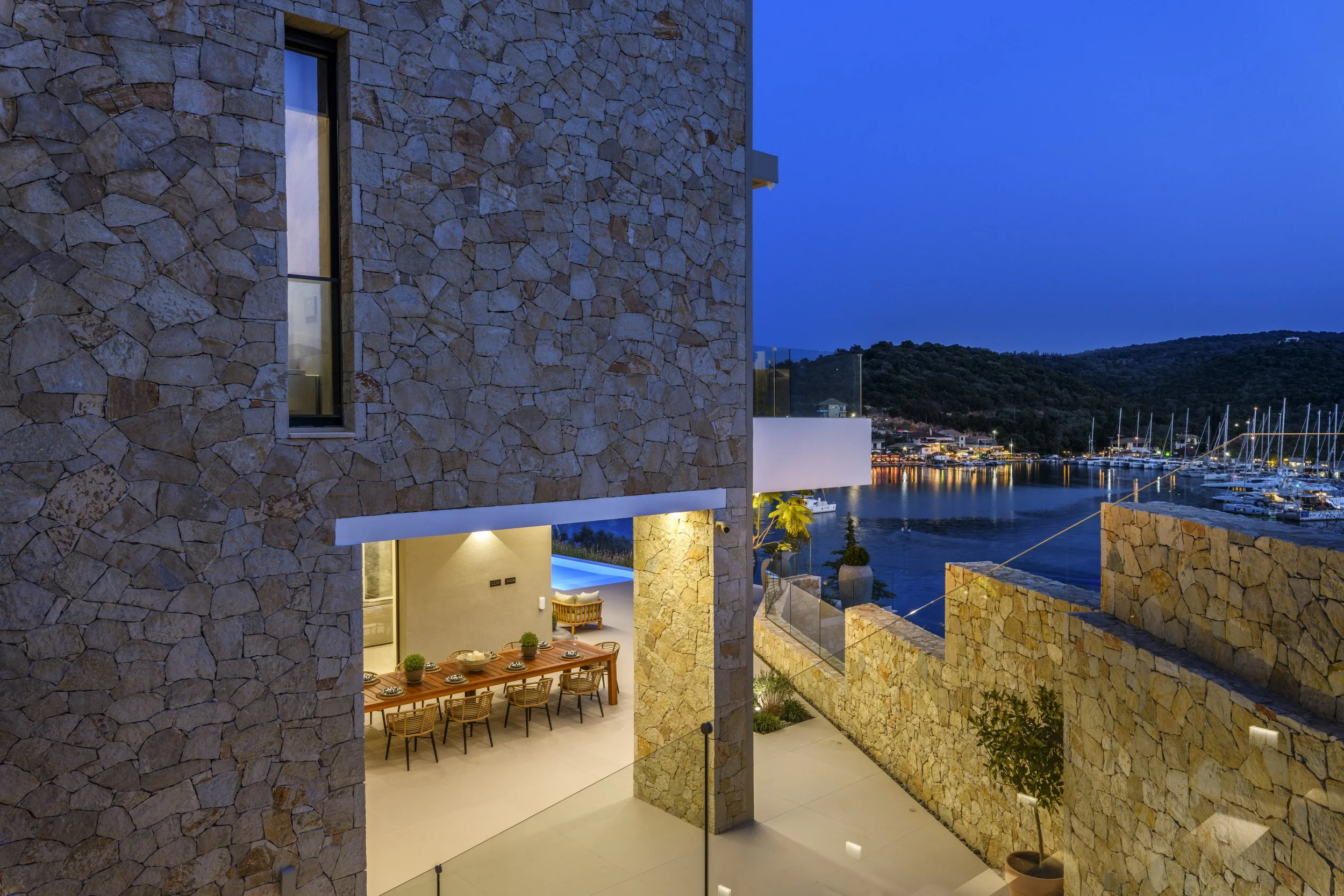Interior view of a modern stone house overlooking a marina with boats, a water body, and hills in the background at dusk. The house has large windows, an outdoor dining area, and stone walls.