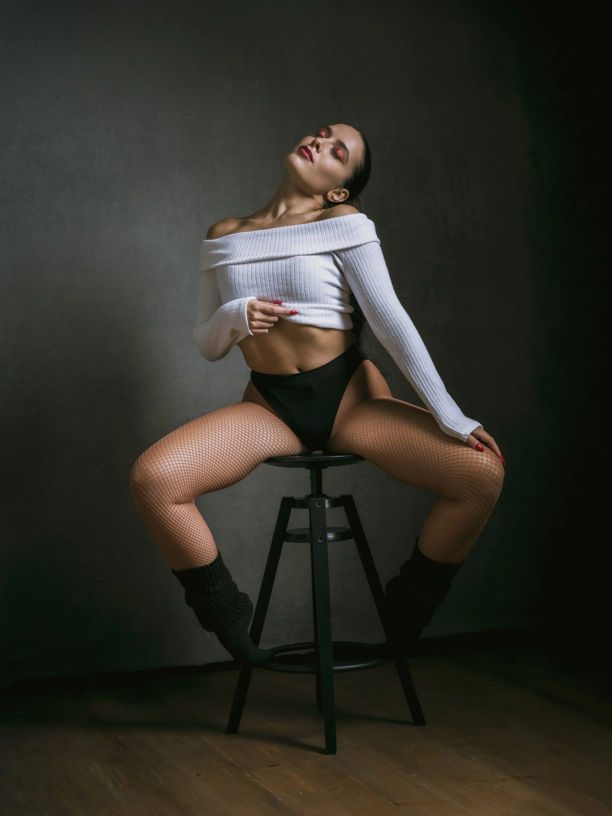 A woman with dark hair and light skin is sitting on a black stool in a dark room with a solid gray background. She is wearing a white off-the-shoulder crop top, black high-waisted underwear, fishnet tights, and black socks. She has her eyes closed, h
